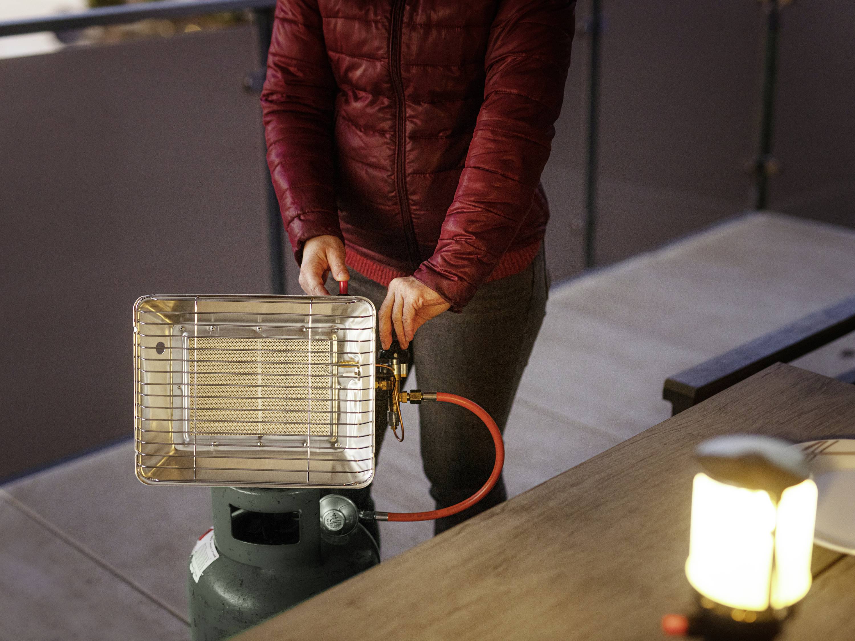 Osoba v červenej bunde pripája prenosný plynový ohrievač na balkóne. V popredí svieti lampa.