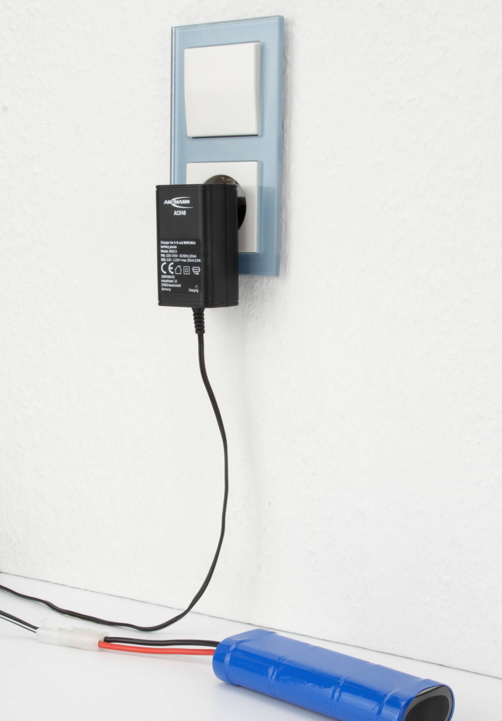 Nabíjačka je zasunutá do elektrickej zásuvky na bielej stene. Kábel vedie k modrému akumulátoru na bielom povrchu.