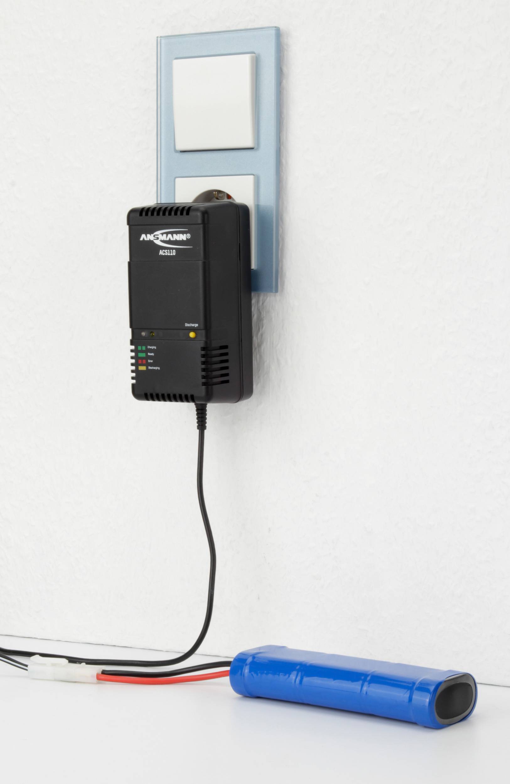 Nabíjačka je zapojená do elektrickej zásuvky a nabíja modrú batériu položenú na stole. Má indikačné svetlá signalizujúce priebeh nabíjania.