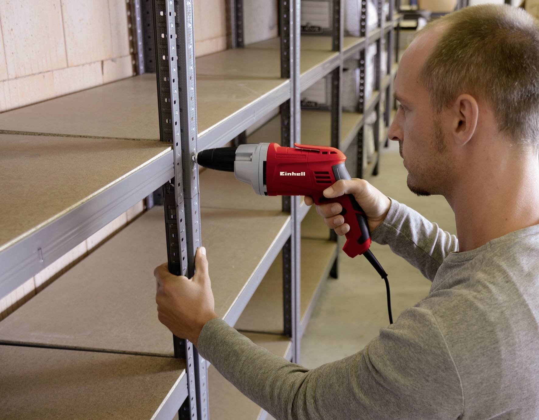 Osoba používa červený vŕtací skrutkovač na montáž kovového regálu v skladovom priestore.