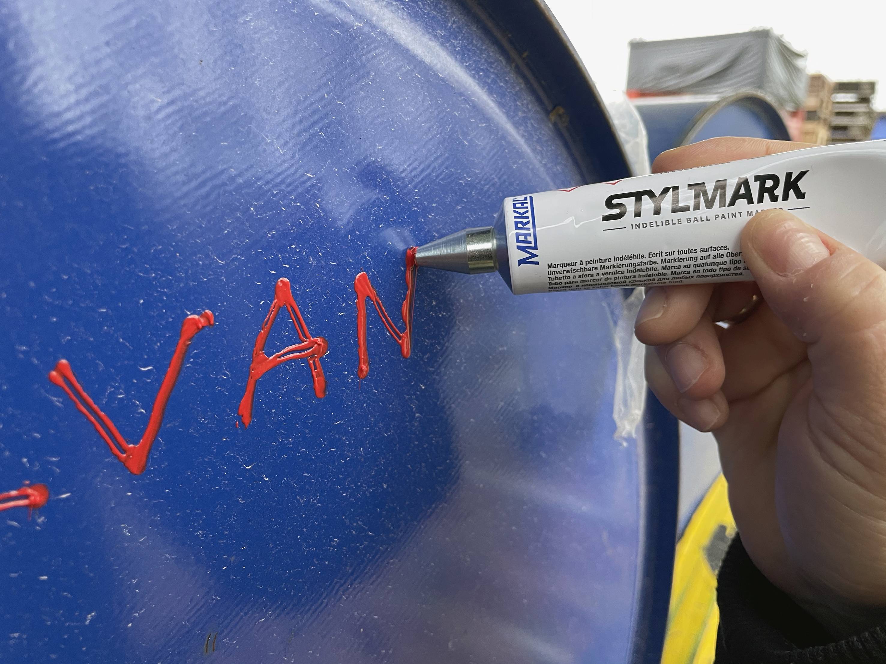 Ruka drží pero a píše 'VAN' červeným písmom na modrom sude.