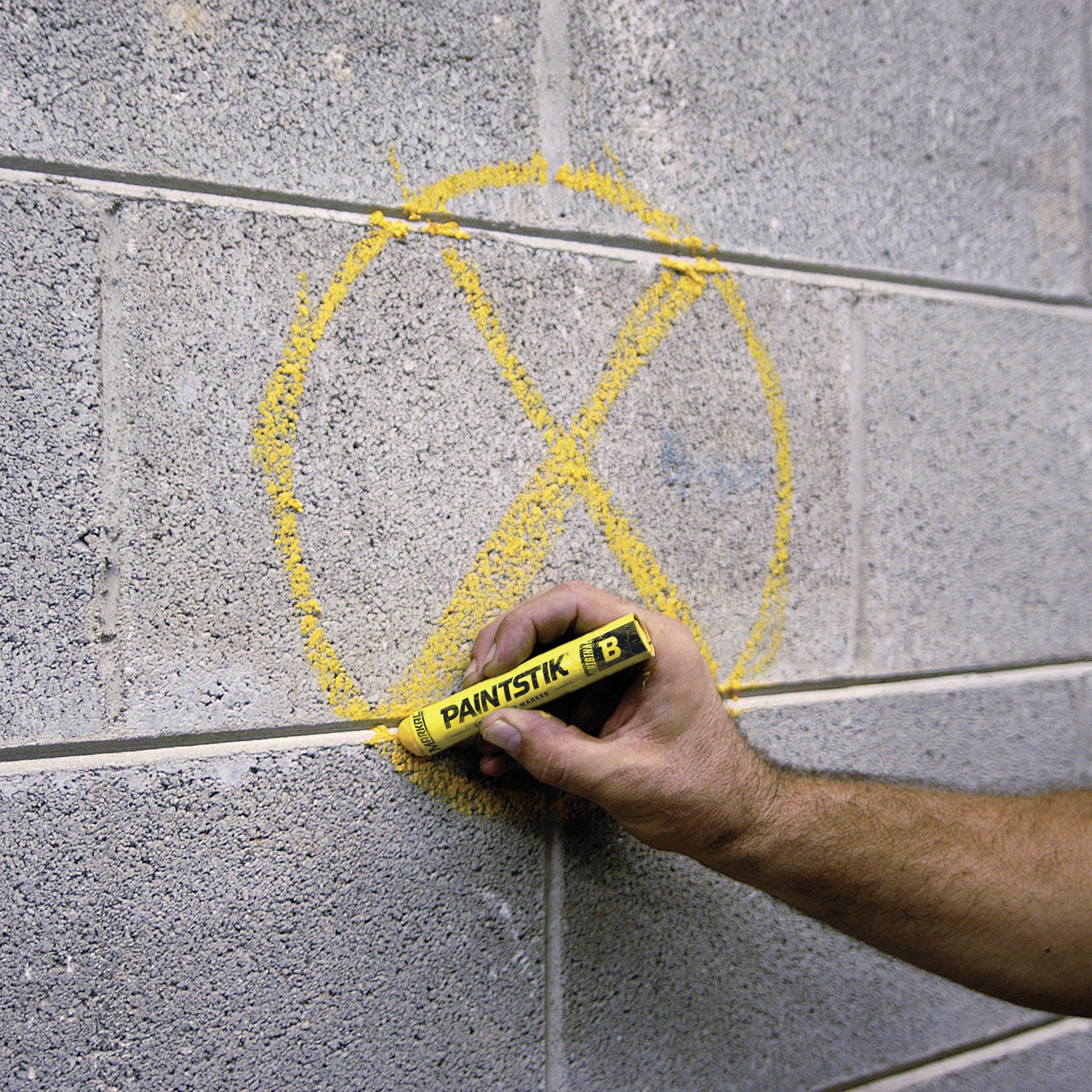 Rameno kreslí žltý krížik na sivú betónovú stenu perom, na ktorom je napísané "Paintstik".