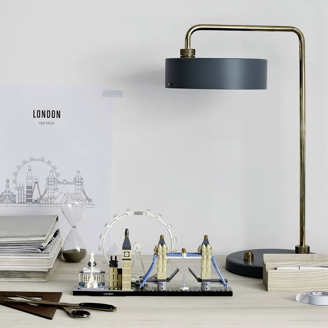 Písací stôl s architektonickým modelom Londýna, stojanová lampa, perá a plány. V pozadí zarámovaný obraz s nápisom 'Londýn'.