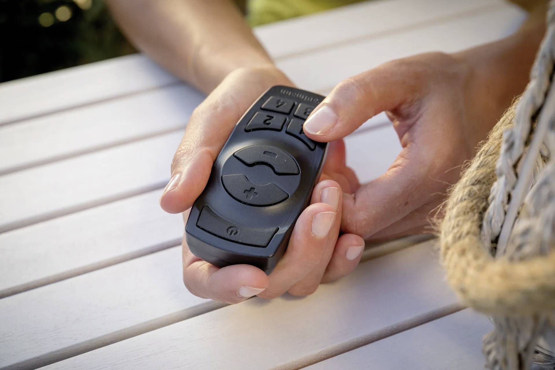 Osoba drží čiernu diaľkový ovládač s tlačidlami. Osoba sedí pri bielom drevenom stole.