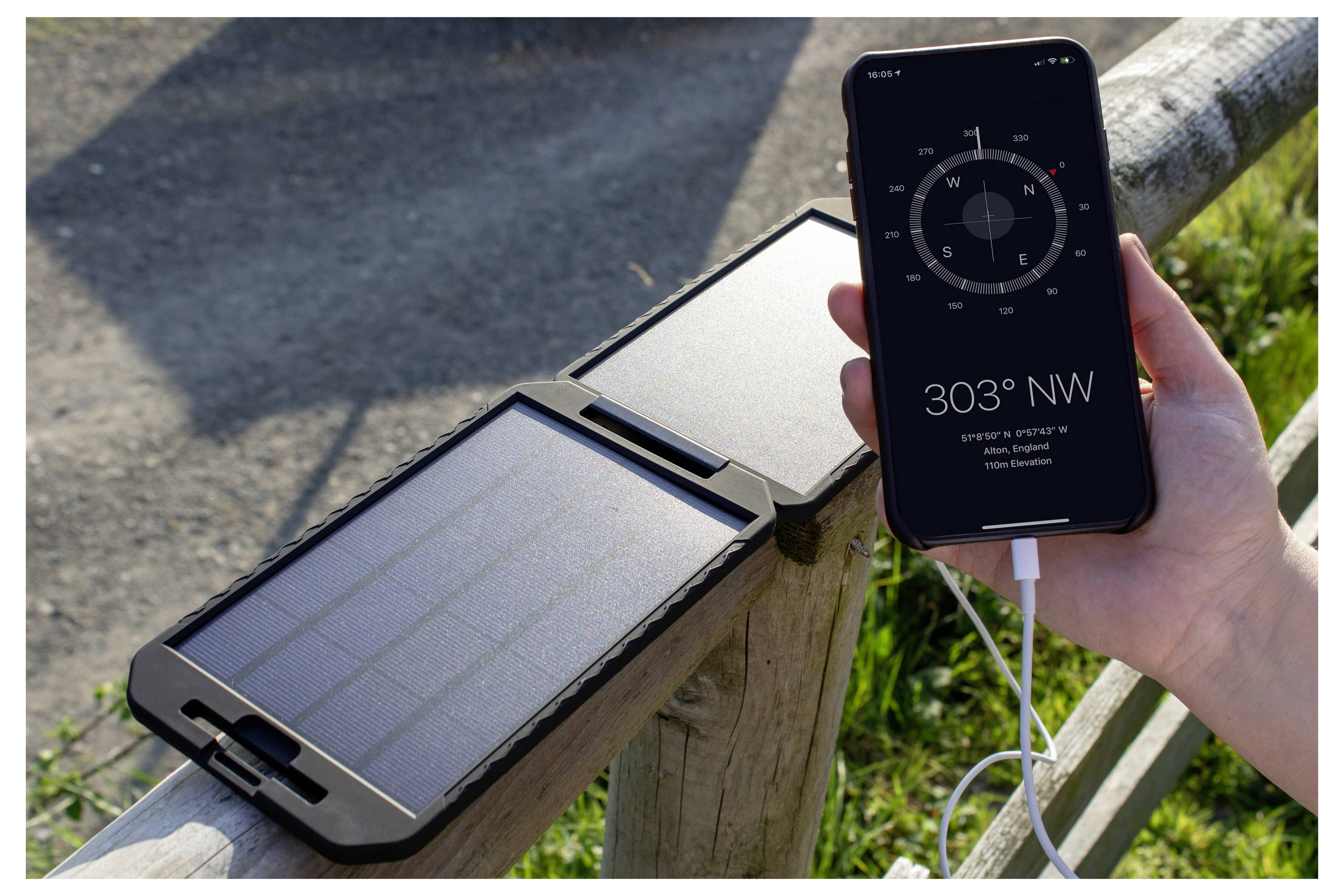 Osoba drží smartfón zobrazujúci kompasovú aplikáciu s údajom „303° SZ", vedľa solárnej nabíjačky na drevenej plot, s pripojeným nabíjacím káblom.