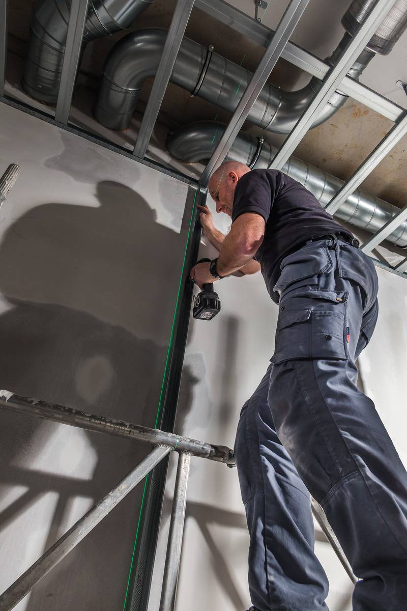 Muž montuje pomocou akumulátorového skrutkovača kovovú konštrukciu na strope. Ventilačné potrubia sú viditeľné.