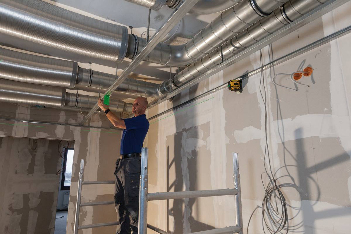 Robotník stojí na lešení a inštaluje ventilačné potrubia na strope nedokončenej časti budovy.