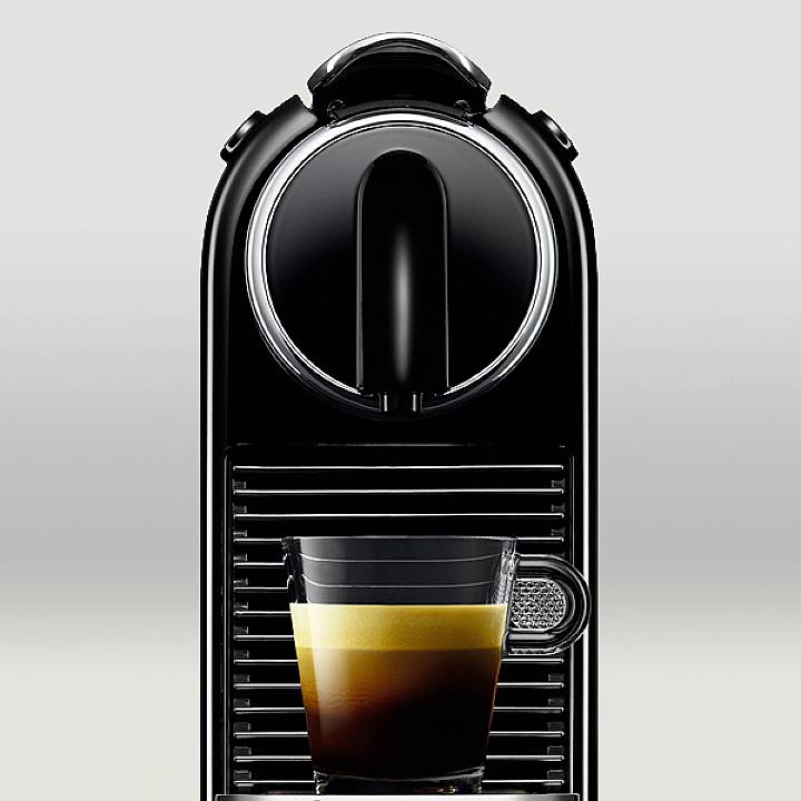 Čierna kávovarka s espresso v priehľadnej šálke, ktorá stojí na odkvapkávacej mriežke.