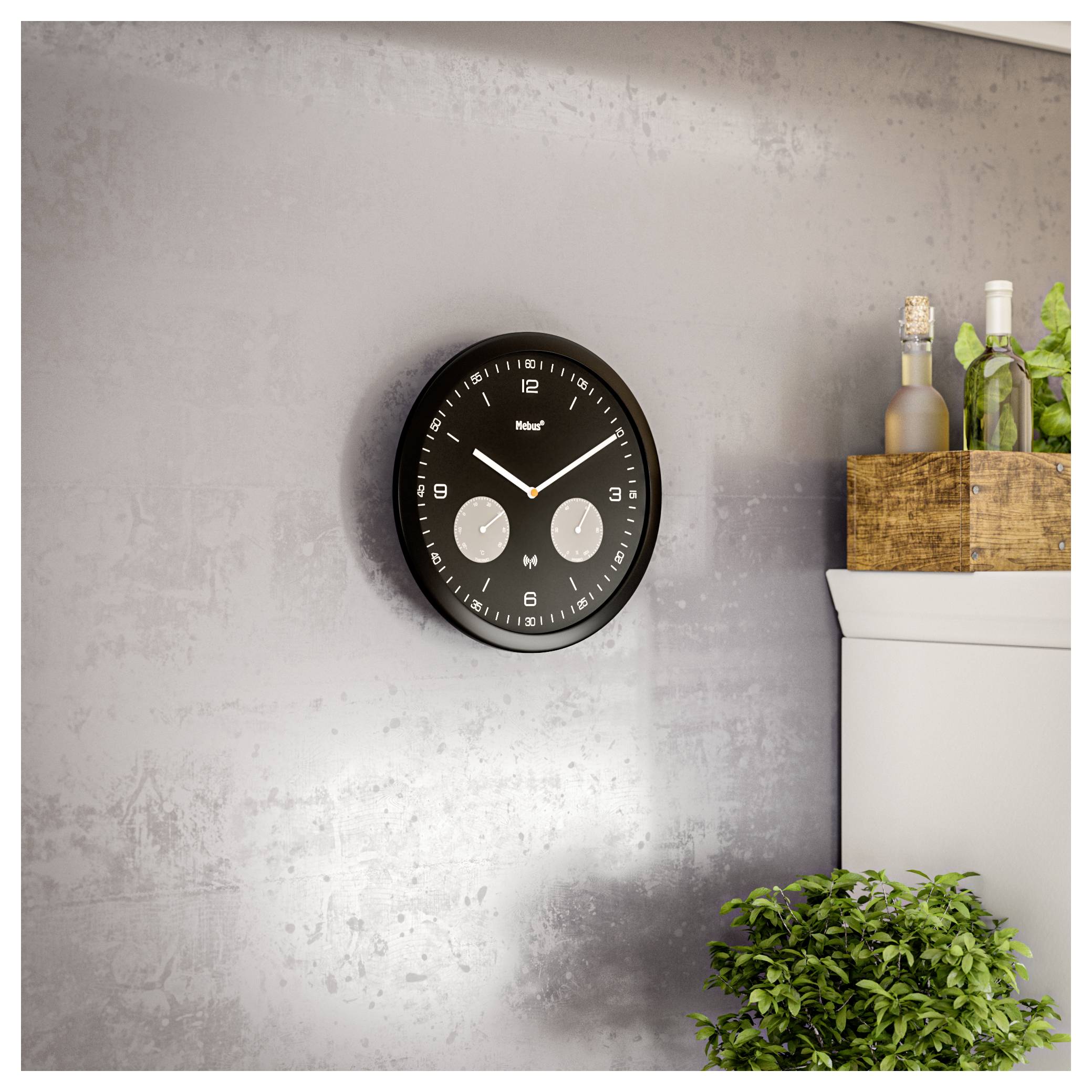 Moderné čierne nástenné hodiny s duálnymi časovými zónami visí na štruktúrovanej sivej stene; neďaleko sú zelené rastliny a drevená krabica s fľašami.