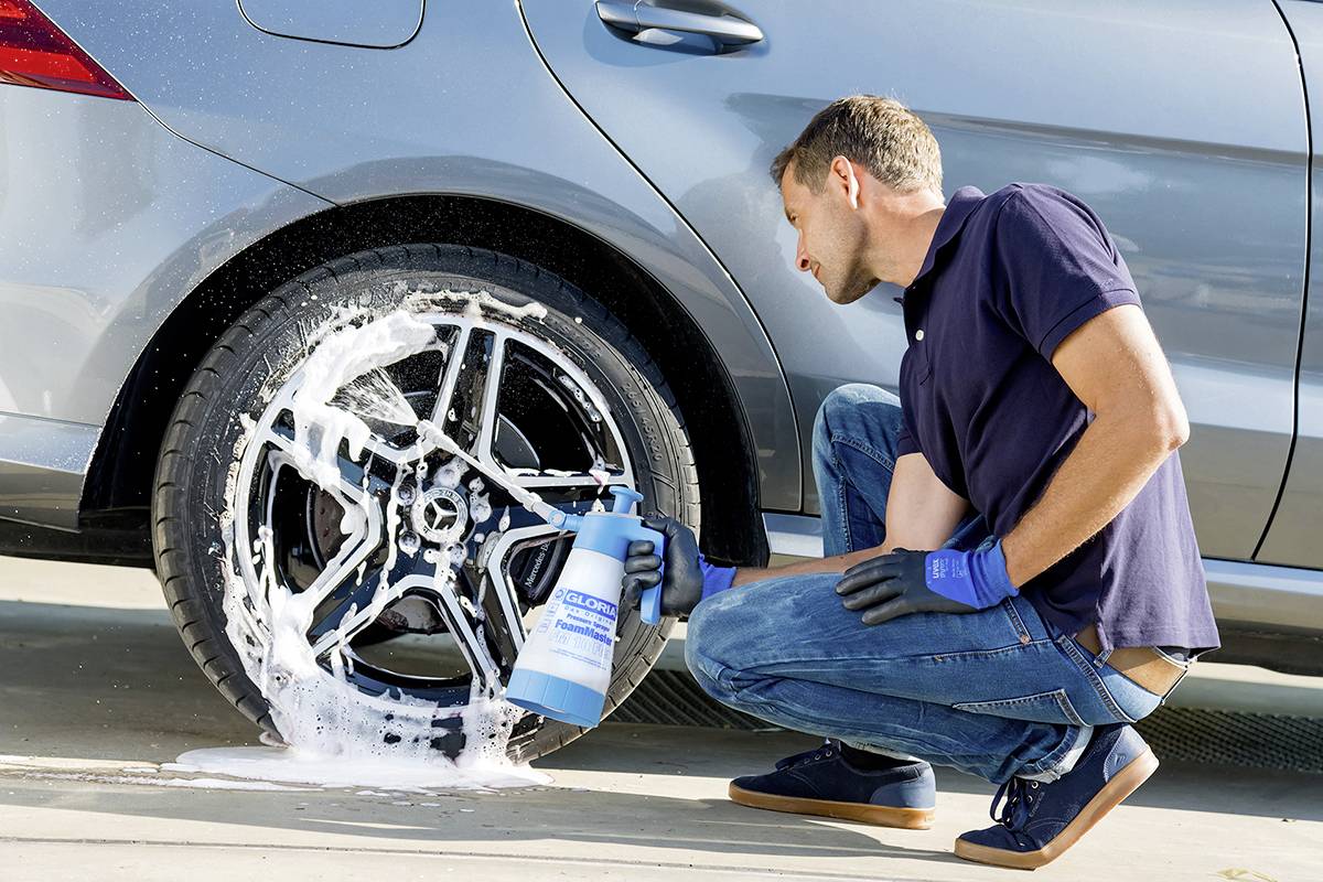 Muž umýva strieborný disk automobilu mydlom, kľačí na zemi, má modré rukavice a strieká čistiaci prostriedok na disk.