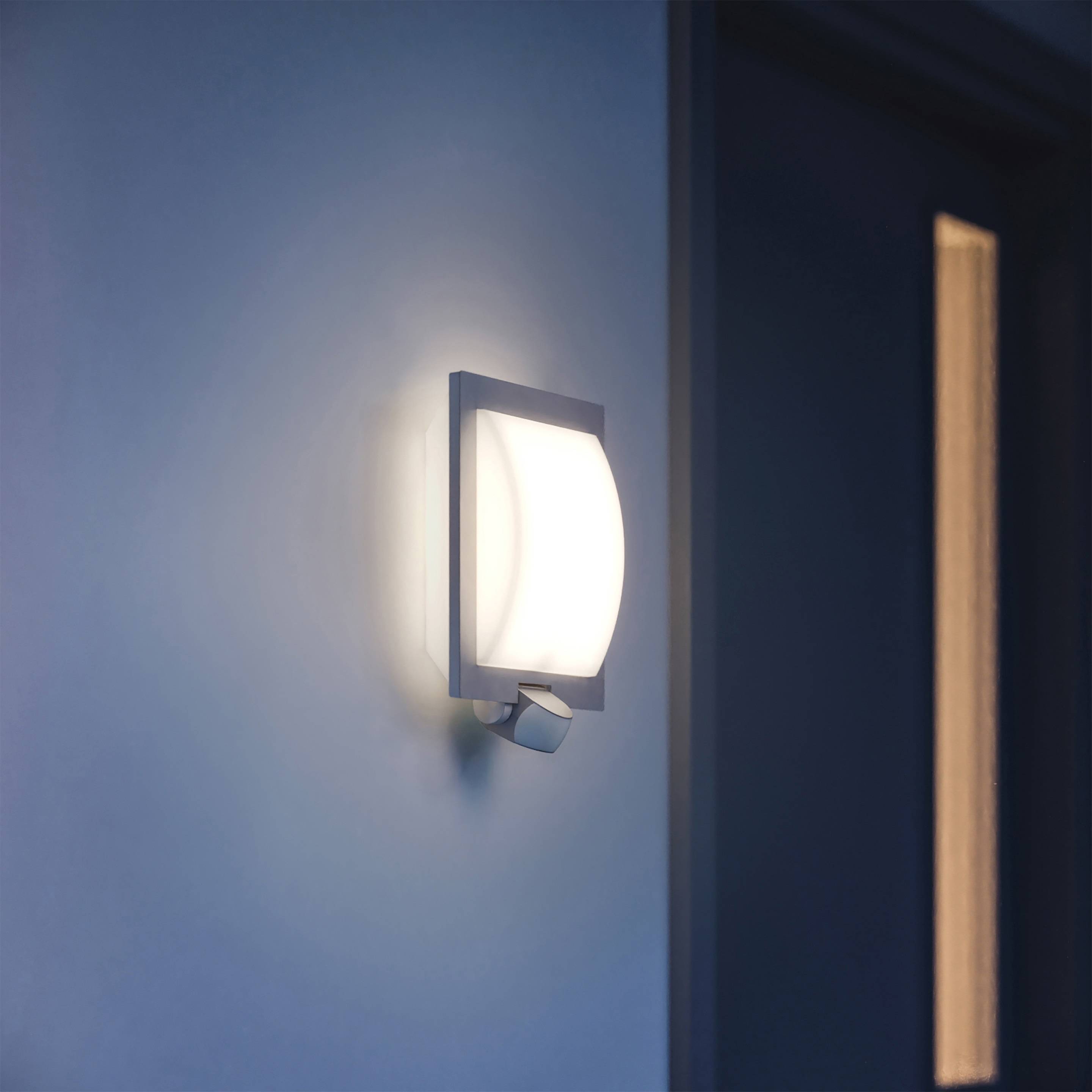 Moderná nástenná lampa svieti vedľa dverí. Lampa má štvorcový dizajn a vyžaruje jemné, teplé svetlo.