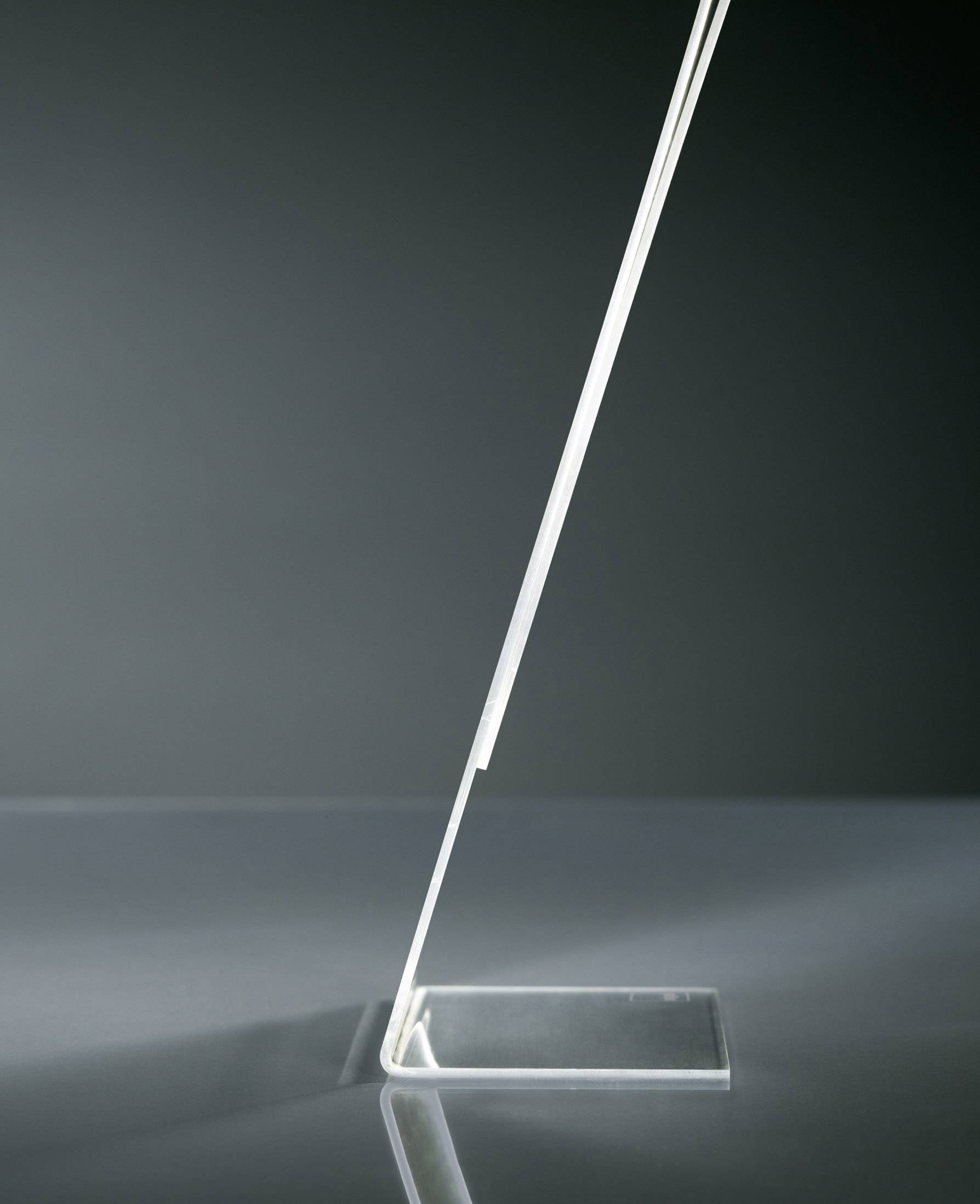 Priehľadný, tenký, šikmý objekt zo skla alebo plastu stojí stabilne na plochom, obdĺžnikovom podstavci.