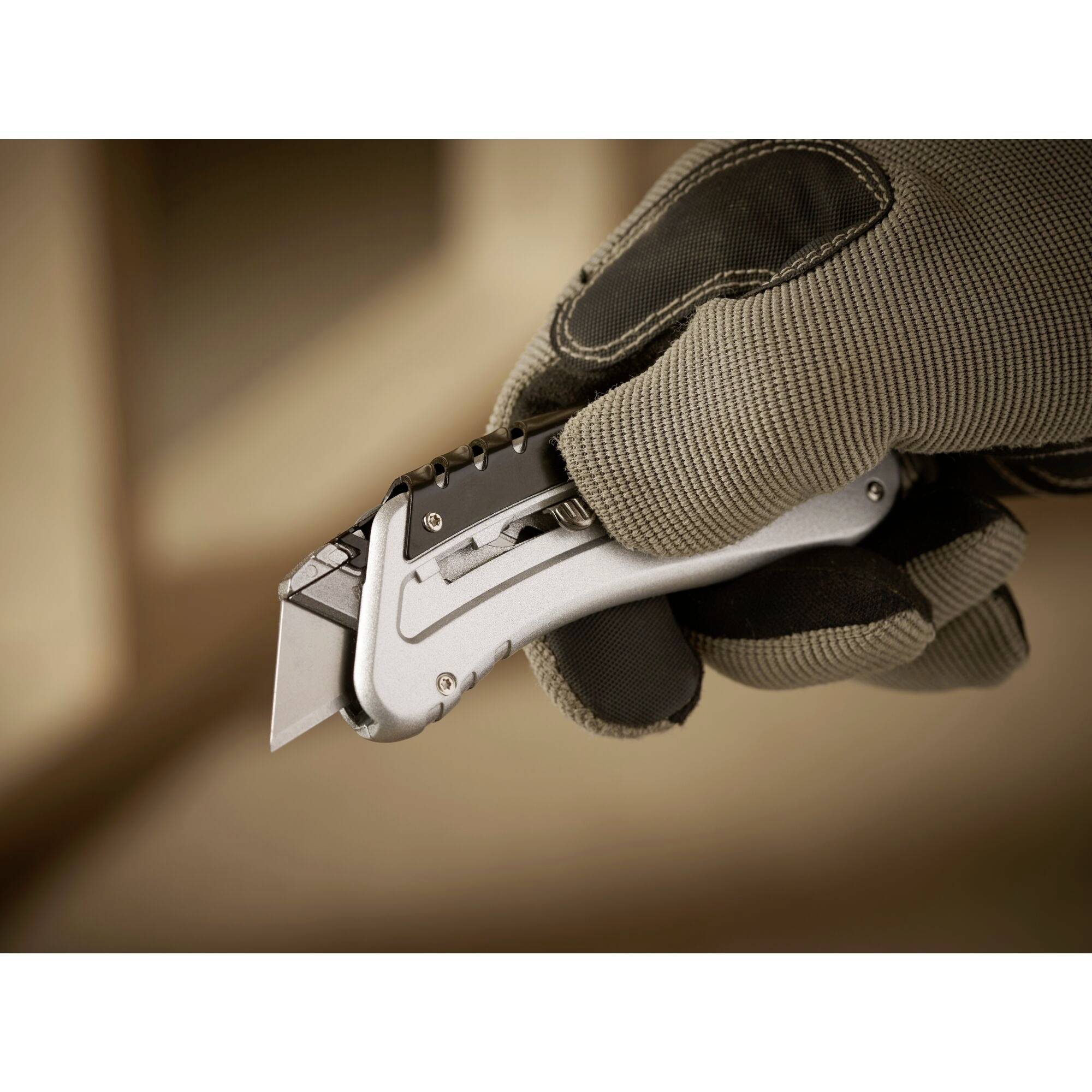 Ruka drží strieborný nôž s čiernou rukoväťou, pripravený na rezanie. Osoba má oblečený hnedý pracovný rukavicu.