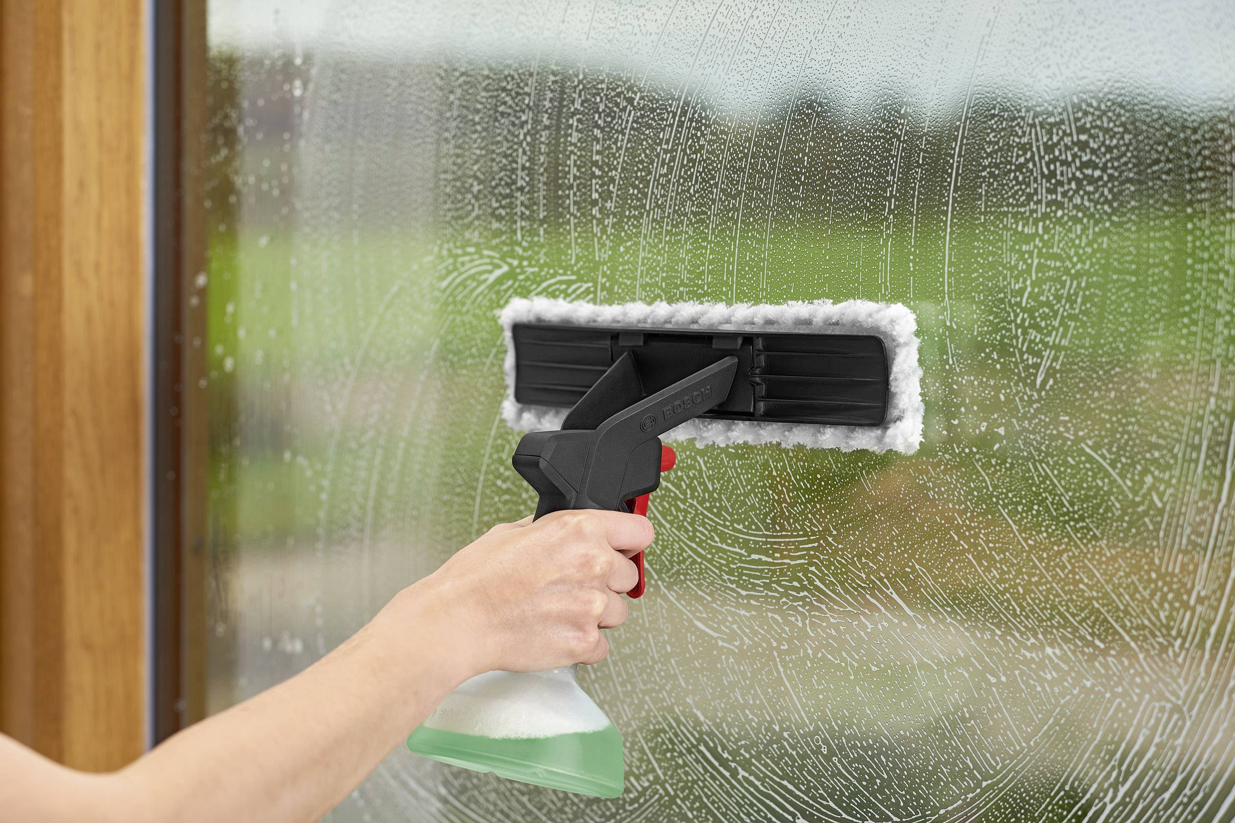 Ruka čistí okno stierkou a rozprašovačom čistiaceho prostriedku. V pozadí je rozmazaná zelená záhrada.