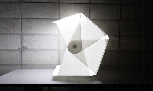 Moderný dizajnový svetelný objekt na stole pred betónovou stenou, vytvorený z geometricky skladaných tvarov vyžarujúcich teplé svetlo.