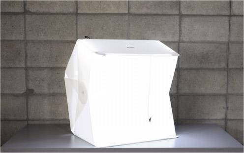 Svetlý fotokocka umiestnený na stole pred sivou tehlovou stenou, používaný na produktovú fotografiu.