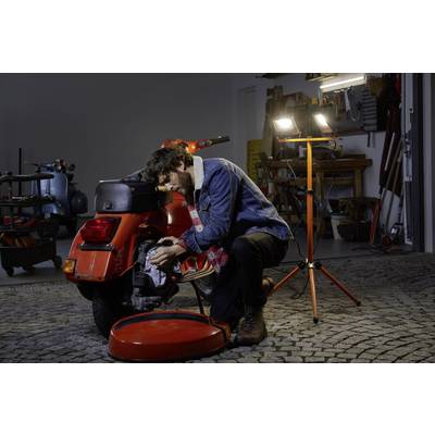 Osoba opravuje oranžovú motorku v garáži. Má na sebe džínsovú bundu a priestor je osvetlený stavebným reflektorom.