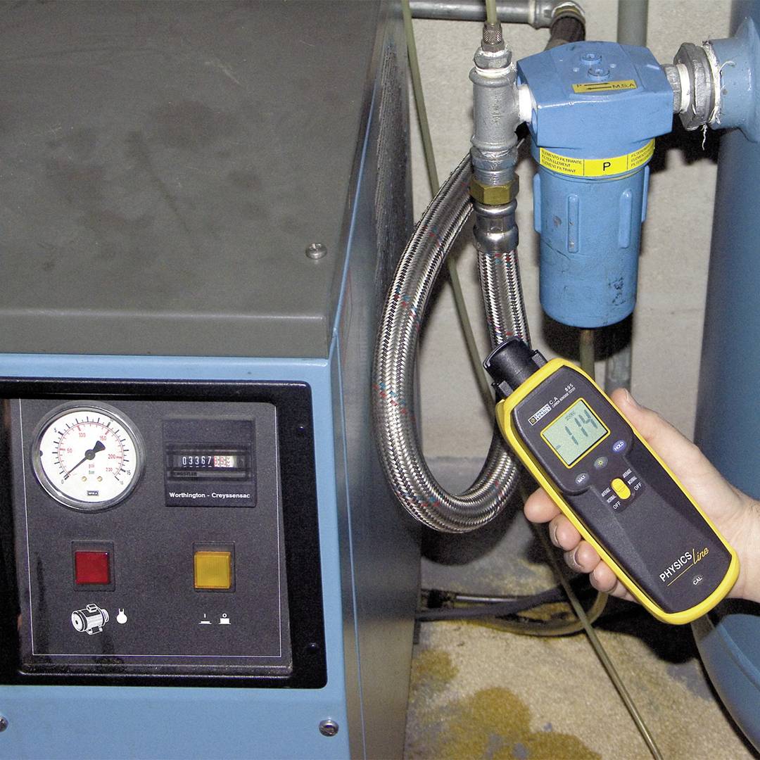 Osoba drží meracie zariadenie pred jednotkou kompresora s tlakovým ukazovateľom a digitálnymi displejmi, aby skontrolovala tlak vzduchu.