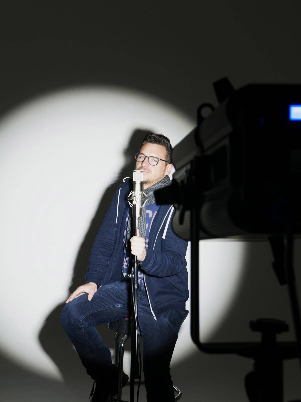 Osoba sedí na stoličke v svetle reflektorov, spieva do mikrofónu. Kamera a vybavenie sú viditeľné v popredí.
