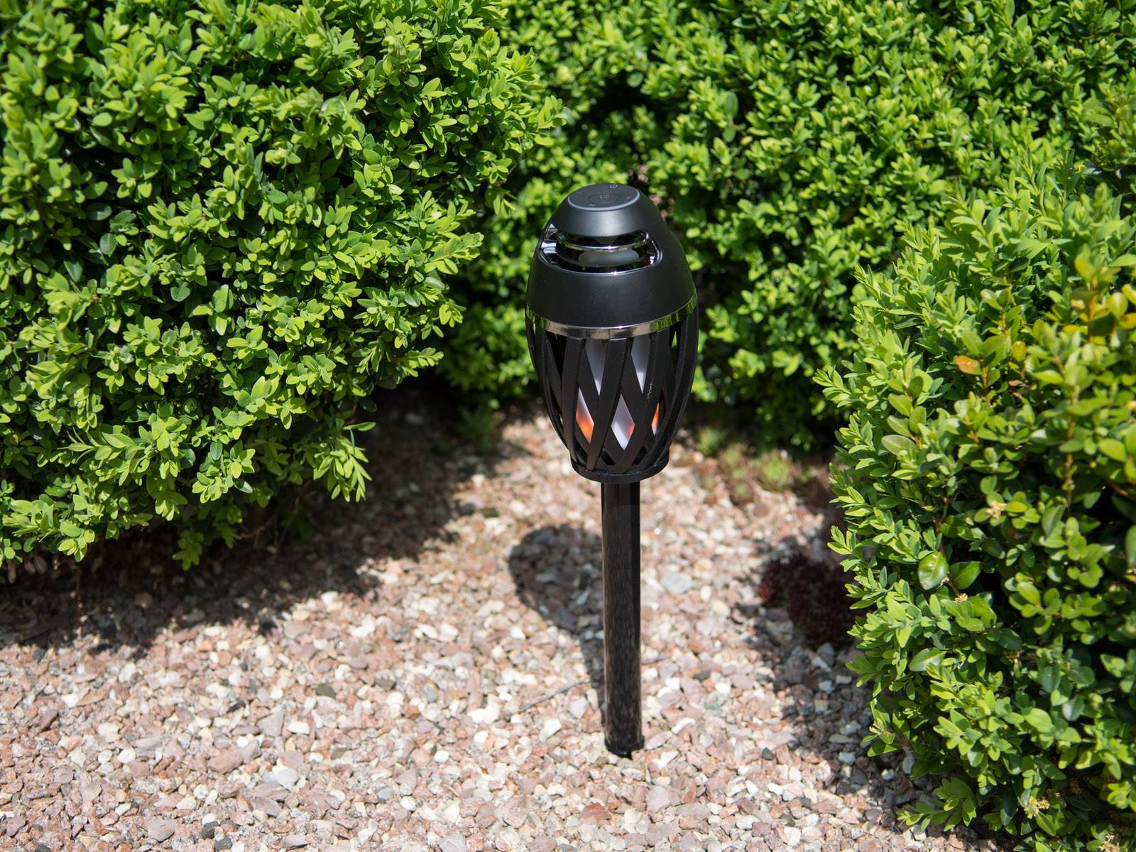 Čierna solárna lampa stojí v záhrade, obklopená zeleným lístím a štrkom. Slúži na osvetlenie a dekoráciu.