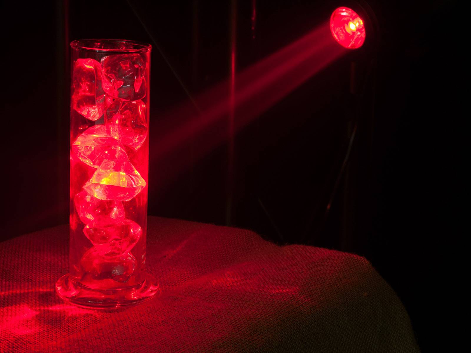 Červený svetelný lúč osvetľuje sklenenný stĺp s dekoratívnymi kameňmi, vytvára odrážajúce a žiariace efekty v tme.