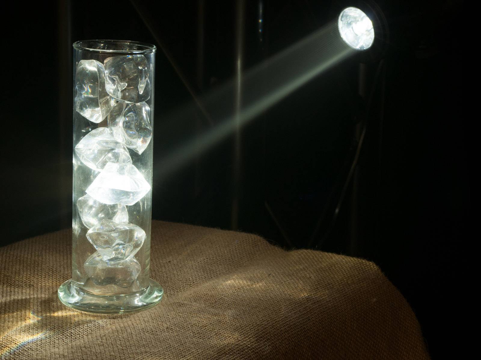 Sklenený valec plný ľadových kociek je osvetlený jasným svetelným lúčom zhora sprava a stojí na hrubej látke.