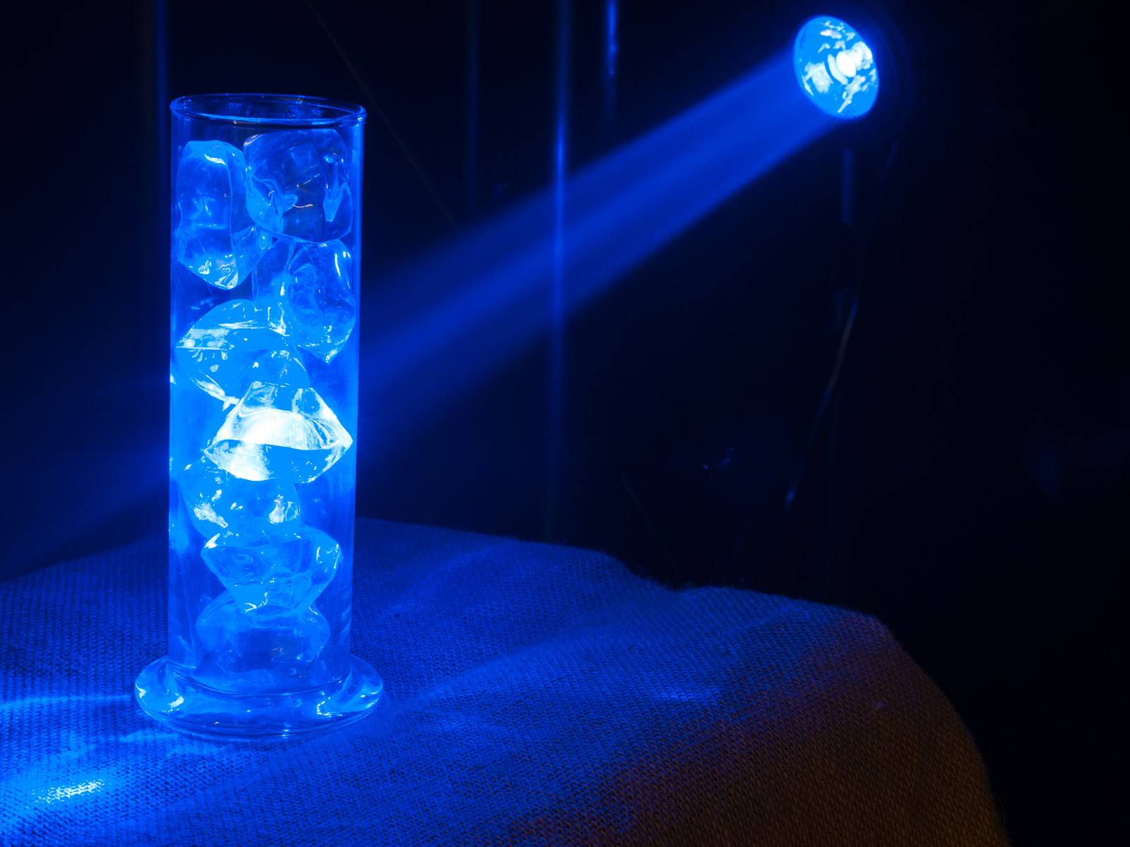 Sklenený valec s ľadovými kockami, osvetlený modrým svetlom, stojí na povrchu potiahnutom látkou a odráža svetlo.