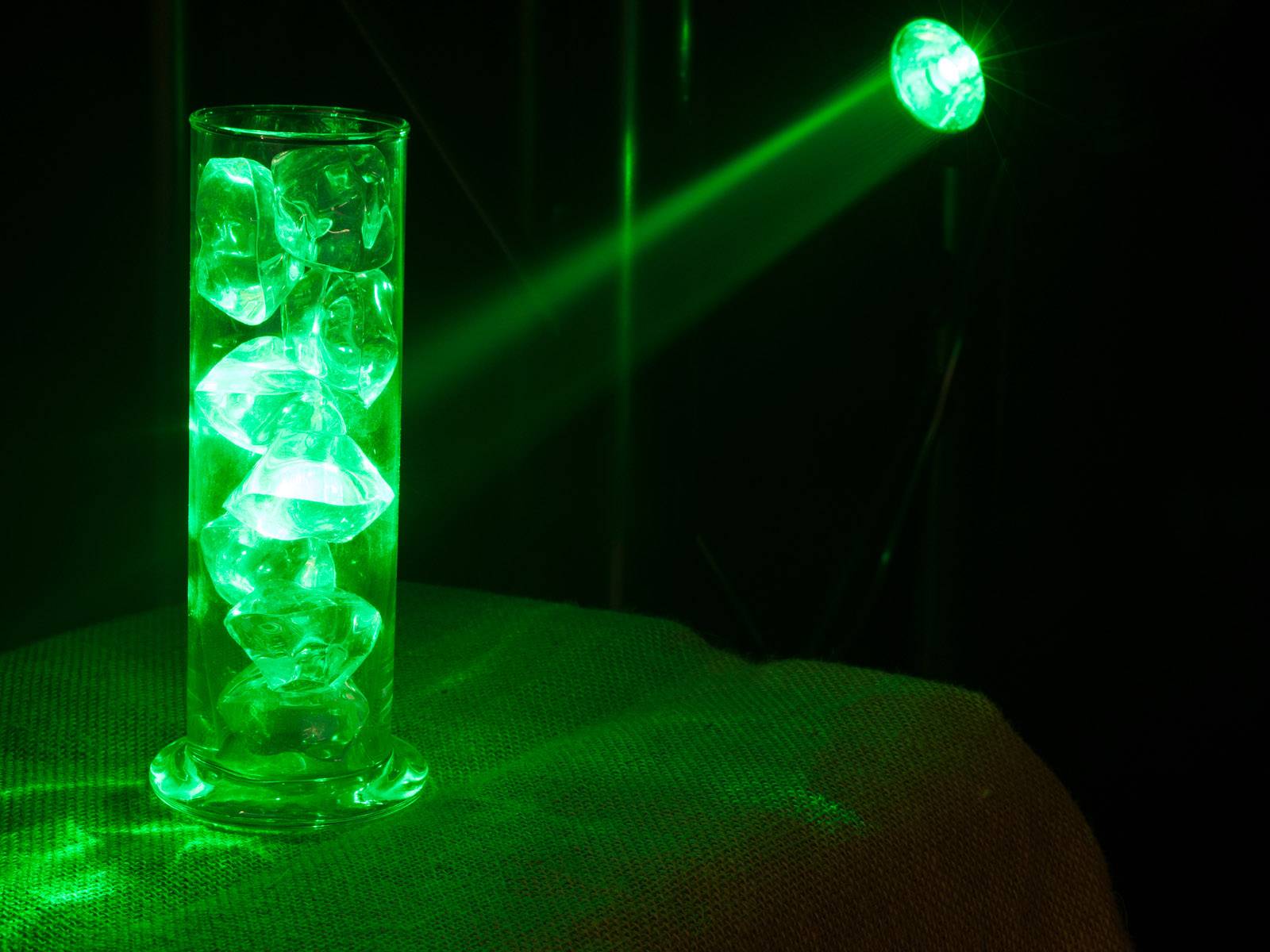 Sklenený valec s ľadovými kockami je osvetlený zeleným svetelným lúčom, vďaka čomu ľadové kocky svietia.