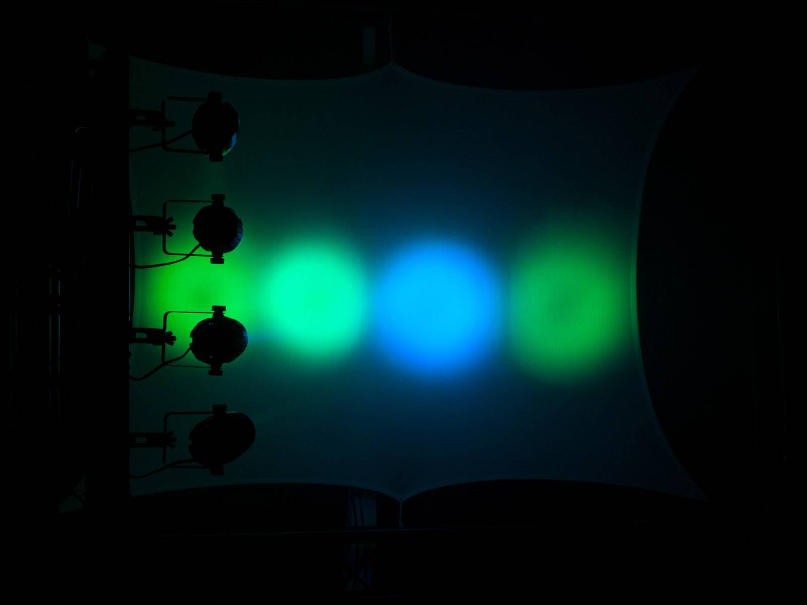 Štyri farebné reflektory premietajú svetelné kruhy v zelenom, modrom a žltom odtieni na tmavú plachtu visiacu v miestnosti.