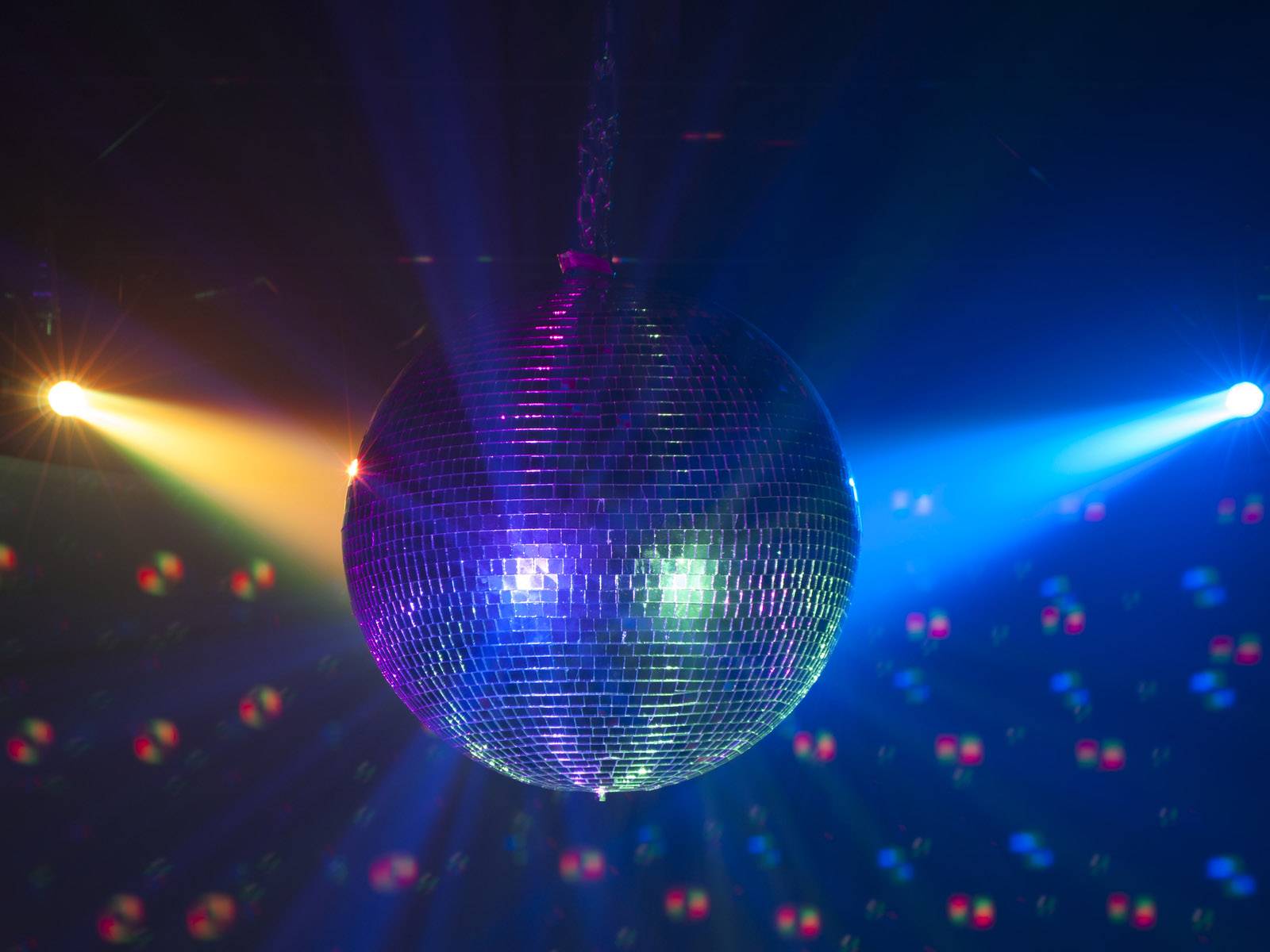 Diskotéková guľa visí v tmavej miestnosti a odráža farebné svetlo zo žltých, modrých a červených reflektorov.