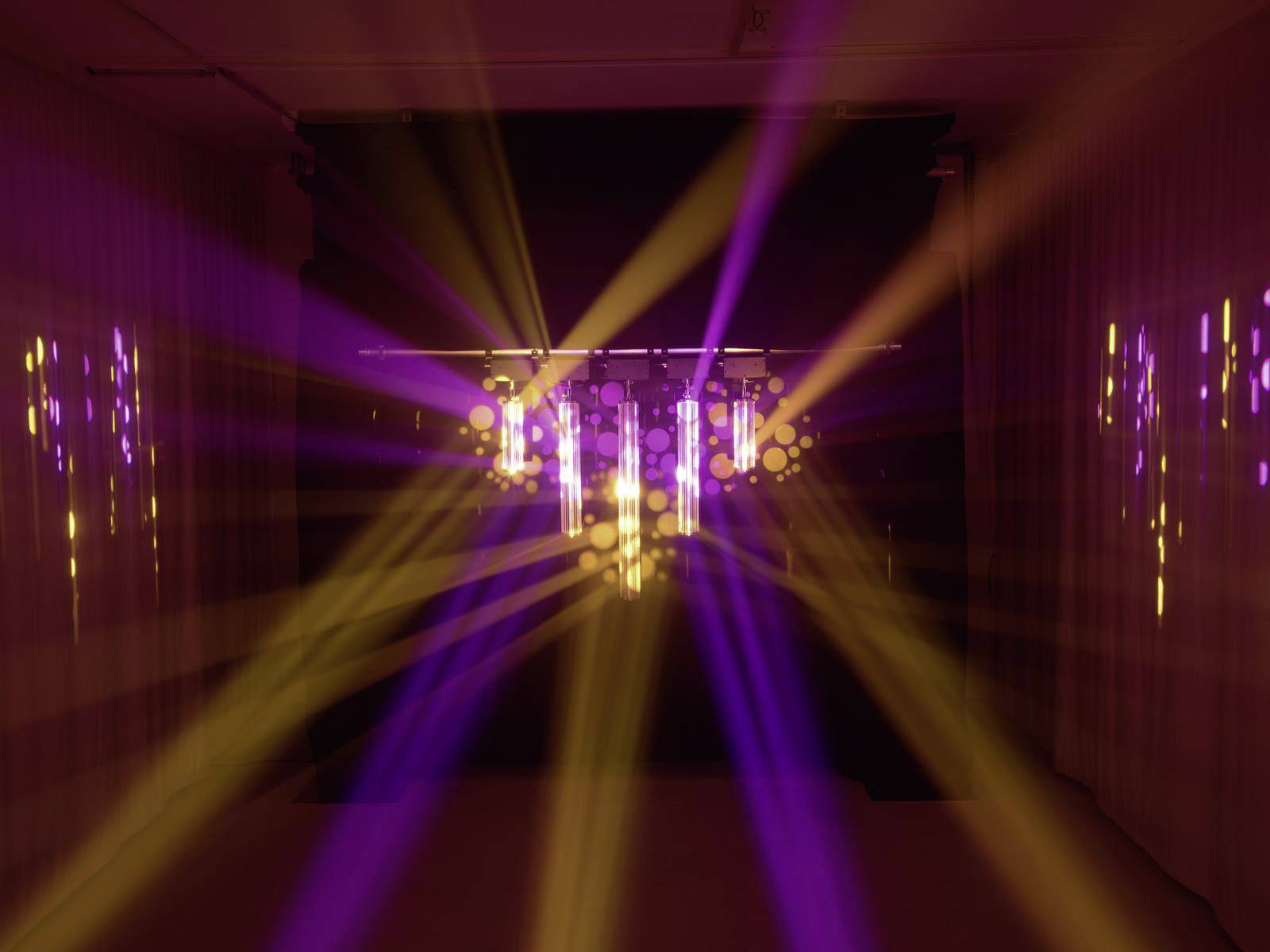 Tmavá miestnosť s farebnými reflektormi, ktoré vytvárajú fialové a žlté svetelné lúče, rozprestierajúce sa rôznymi smermi.