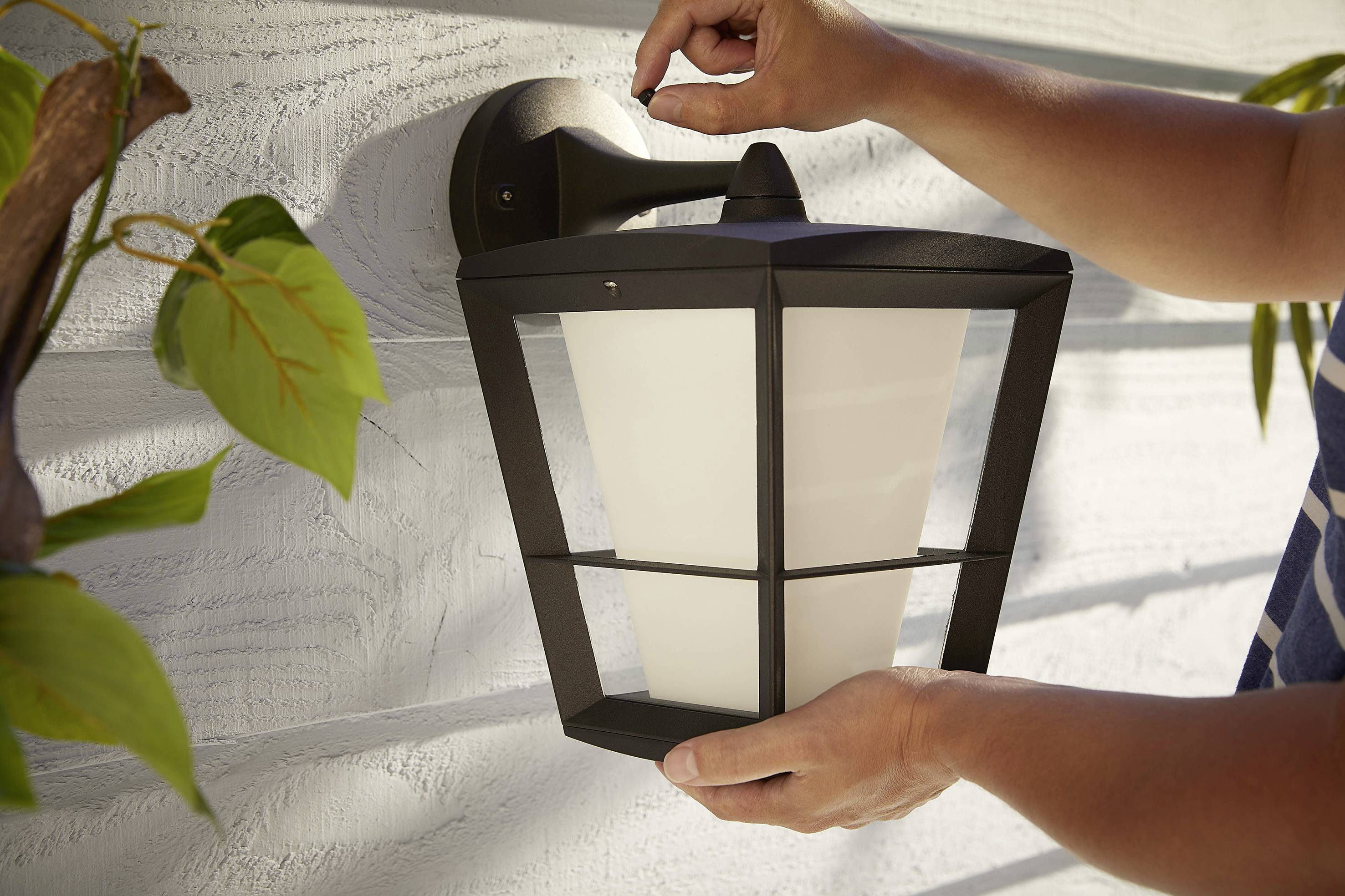 Osoba montuje čiernu vonkajšiu svetelnú lampu na svetlej stene, obklopená zelenými listami.
