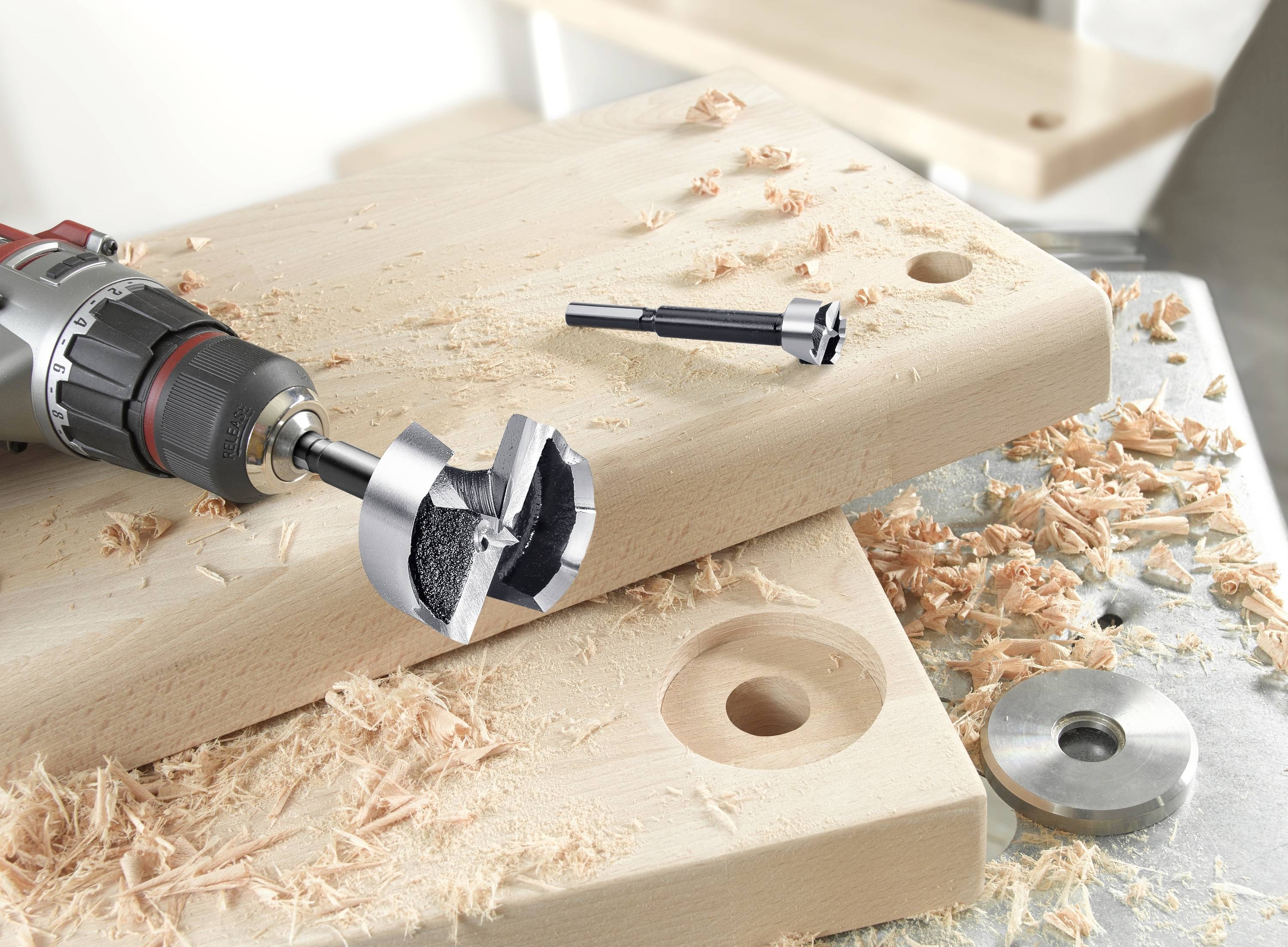 Vŕtačka s Forstnerovým vrtákom na drevenej doske, triesky obklopujú pracovný priestor, zobrazuje scénu opracovania dreva.