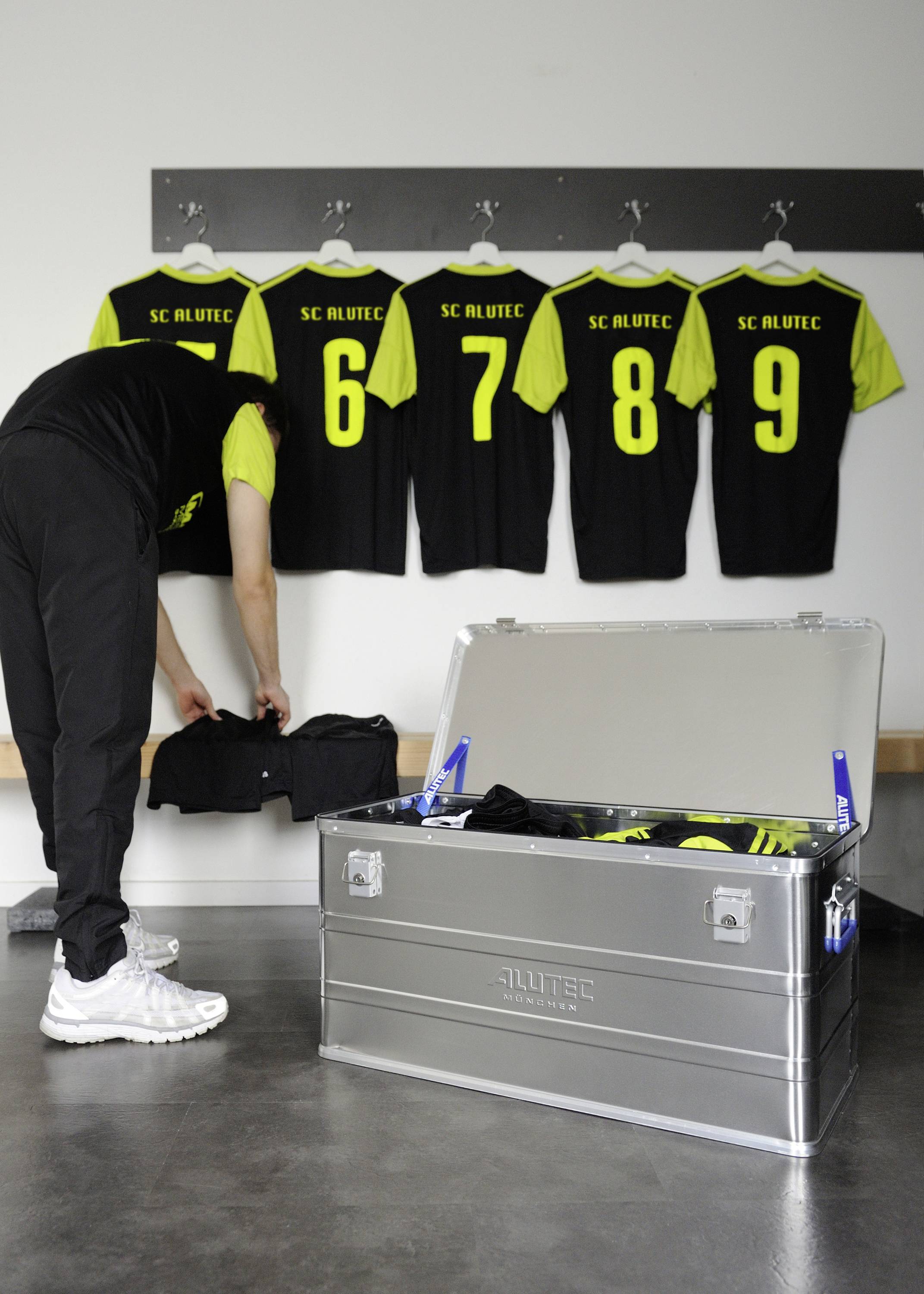 Osoba vyberá futbalové dresy zo striebornej debny v šatni; v pozadí visia číslované dresy na vešiakoch.