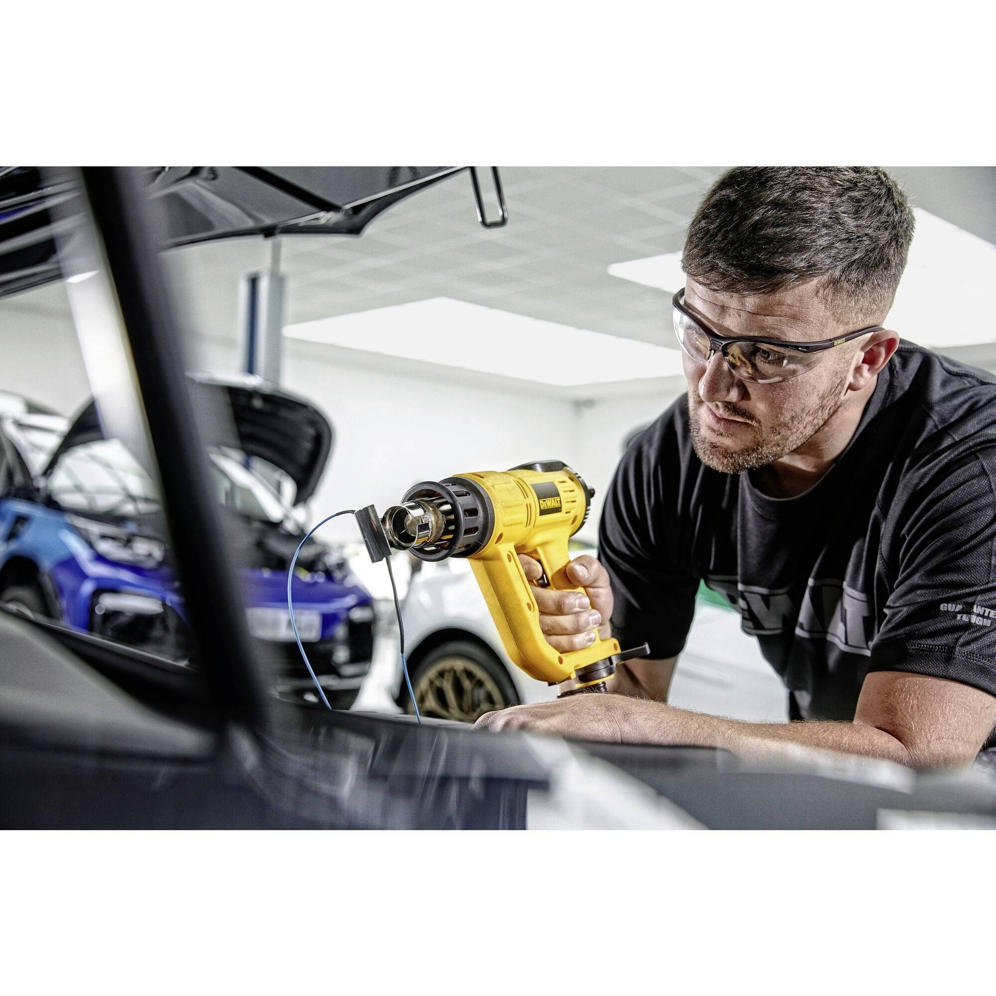 Muž v ochranných okuliaroch používa žltý teplovzdušný fén v dielni. V rozmazanom pozadí sú viditeľné autá.