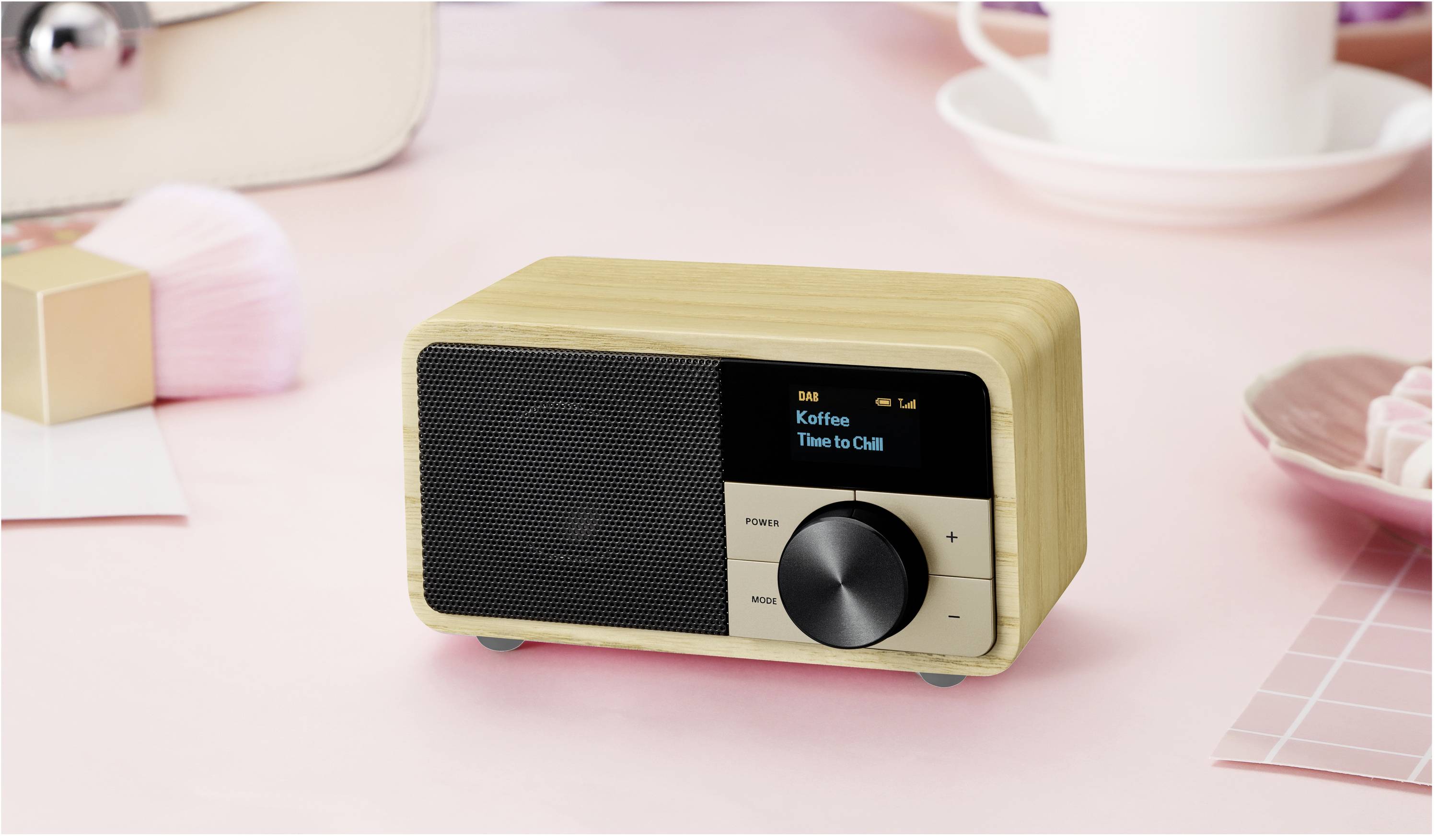 Malé drevené rádio stojí na ružovom stole. Na displeji svieti nápisy 'Káva' a 'Čas na oddych'. Vedľa neho stojí šálka.