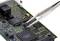 Pinzeta drží malý čierny čip nad zelenou doskou plošných spojov s početným elektronickým príslušenstvom.