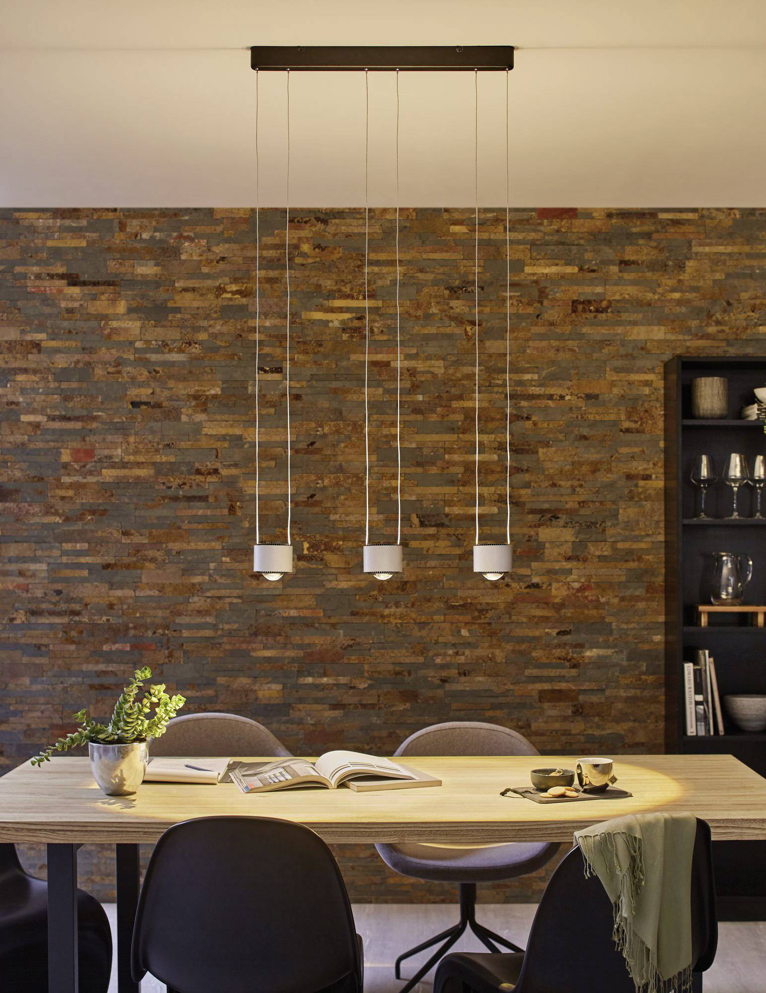Jedálenská oblasť s dreveným stolom, štyrmi stoličkami a knižnicou. Moderné závesné lampy nad stolom. Teplá tehlová stena v pozadí.