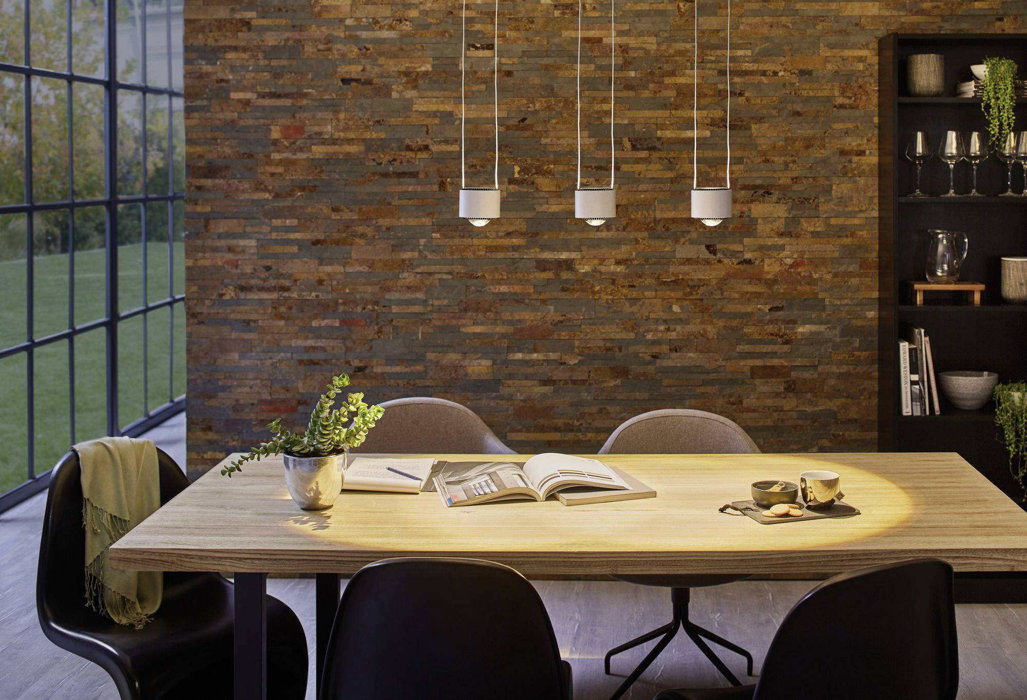 Jedáleň s dreveným stolom, piatimi stoličkami a knihami na stole. Tri lampy visia nad stolom. Veľké okná ponúkajú výhľad na záhradu.