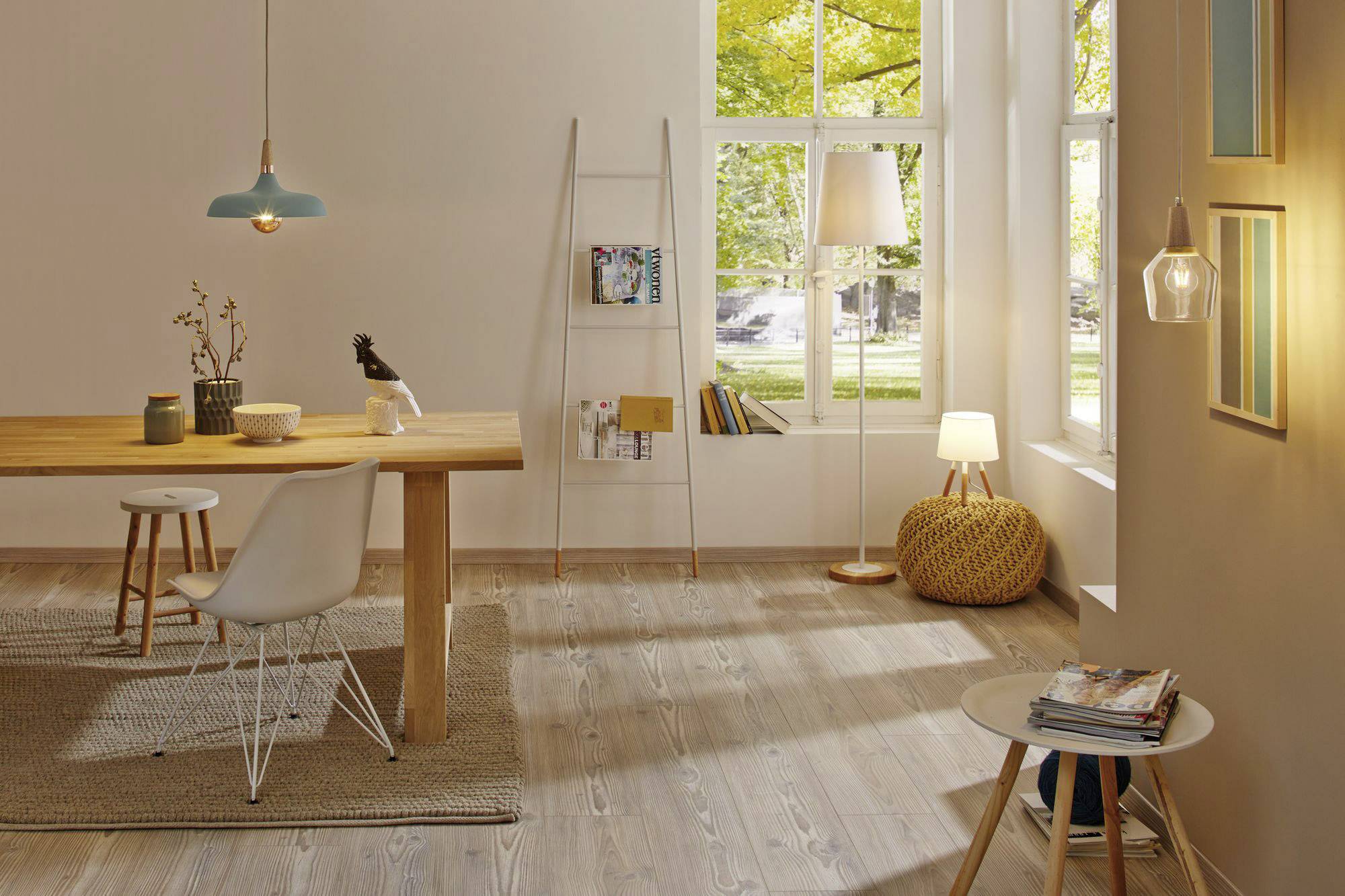 Svetlá obývačka s dreveným stolom a stoličkami, knižnicou, kobercom a stojanými lampami. Veľké okná púšťajú dovnútra denné svetlo.