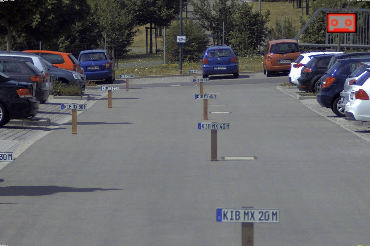 Parkovisko s vyhradenými parkovacími miestami, označenými stĺpikmi s nápismi. V pozadí sú stromy a ďalšie autá.