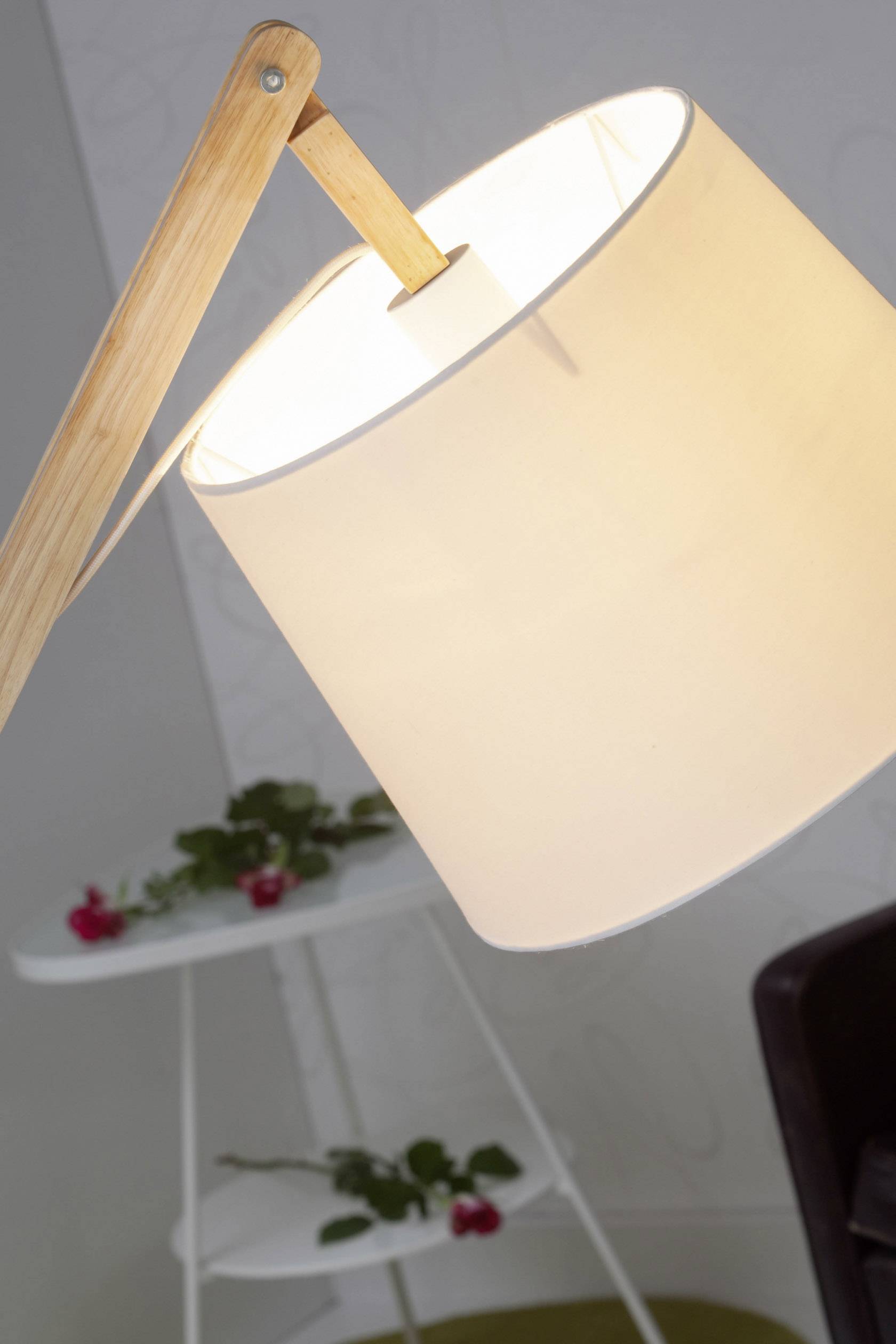Stojaca lampa naklonená do strany so svetlým, okrúhlym tienidlom a dreveným stojanom, umiestnená pred stolom s červenými kvetmi ako dekoráciou.