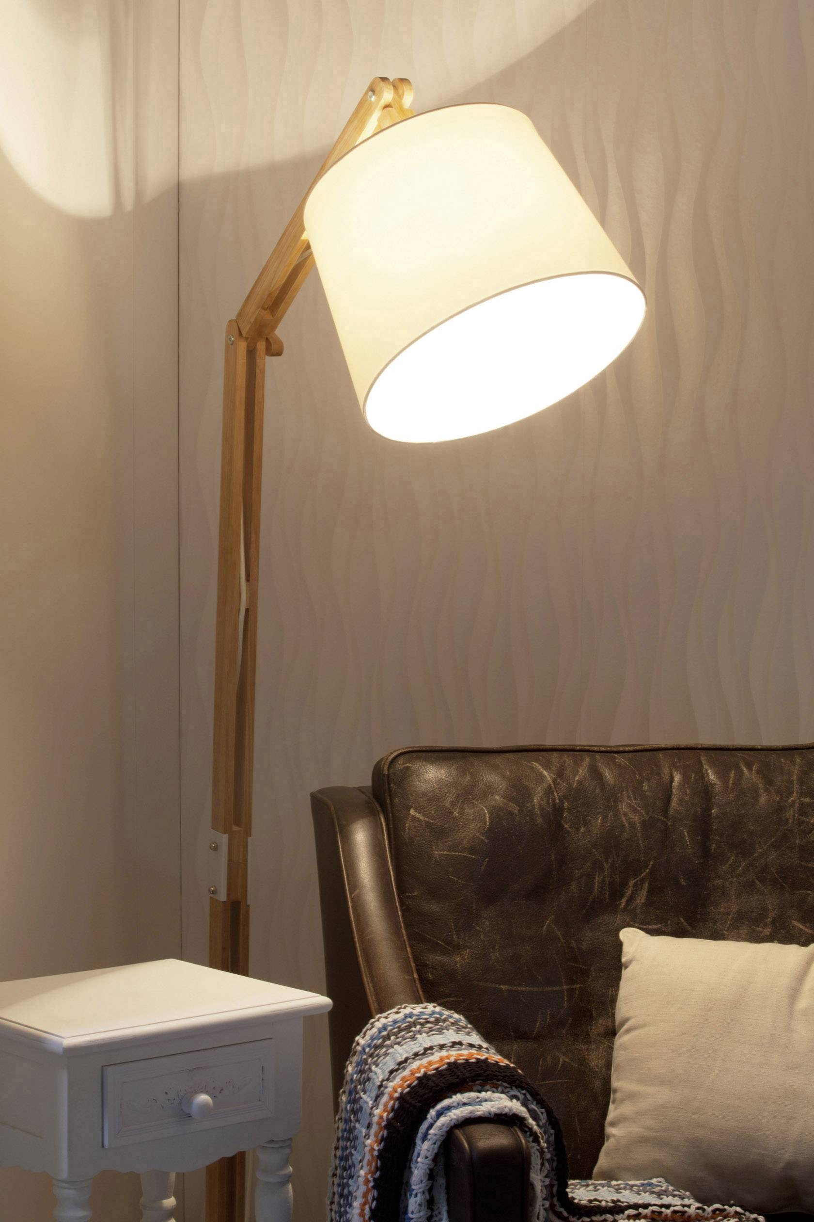 Stojacia lampa vedľa hnedého kožného kresla s vankúšom a háčkovanou dekou, vedľa ktorej stojí malý biely príručný stolík.