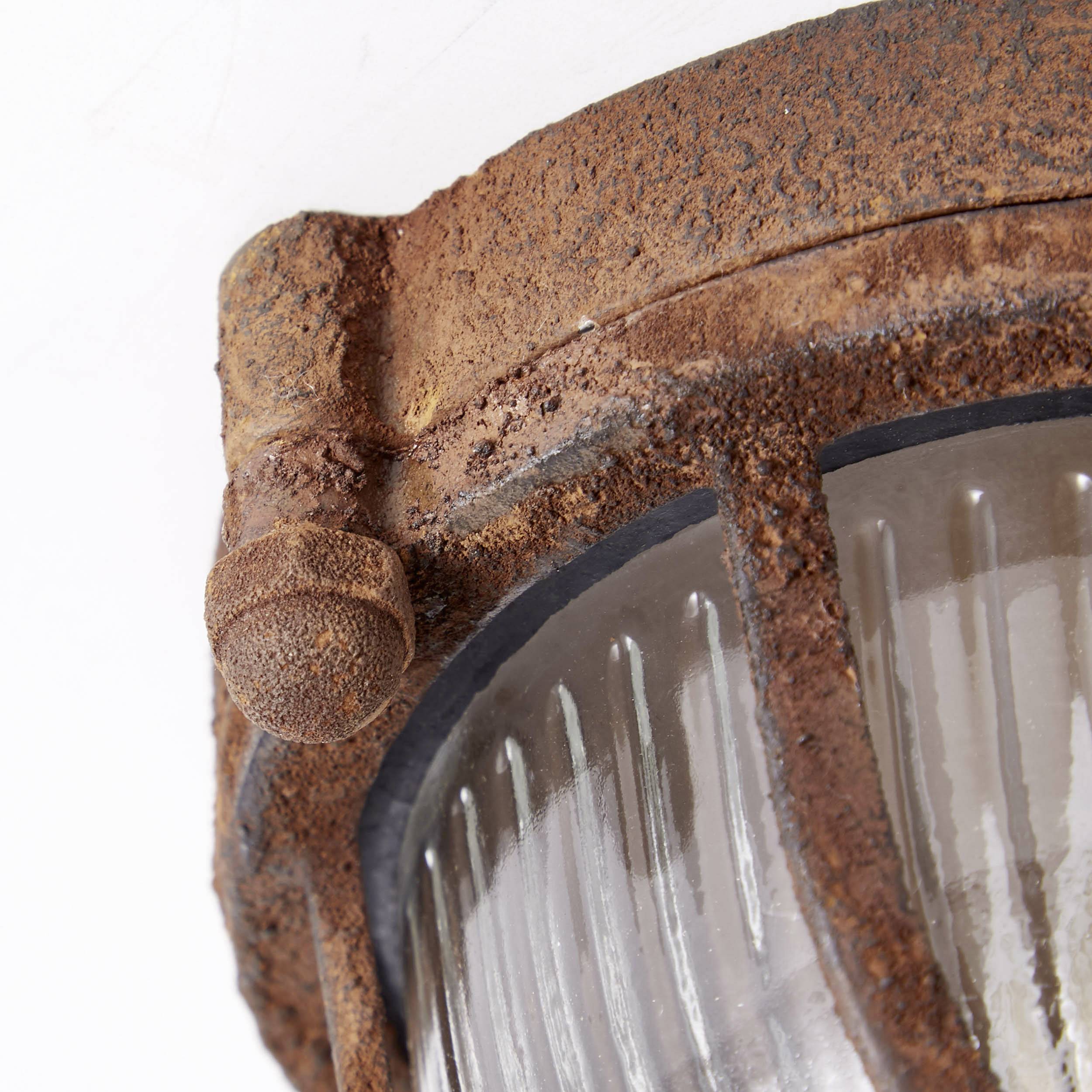 Hrdzavá vonkajšia lampa so skleneným krytom, ktorá vykazuje viditeľné známky opotrebenia.