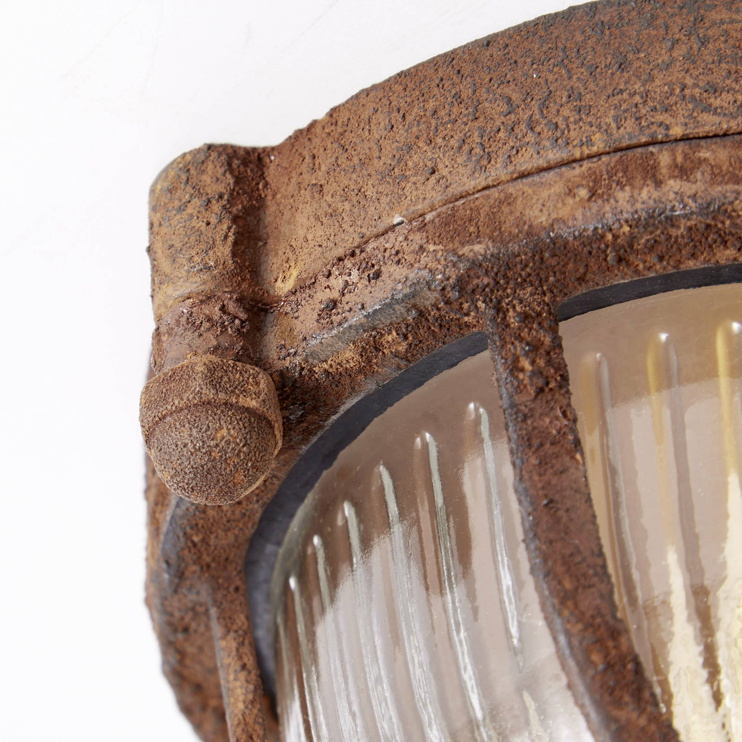 Detailný záber starej, hrdzavej kovovej lampy so skleneným tienidlom. Povrch vykazuje výrazné známky opotrebenia a korózie.