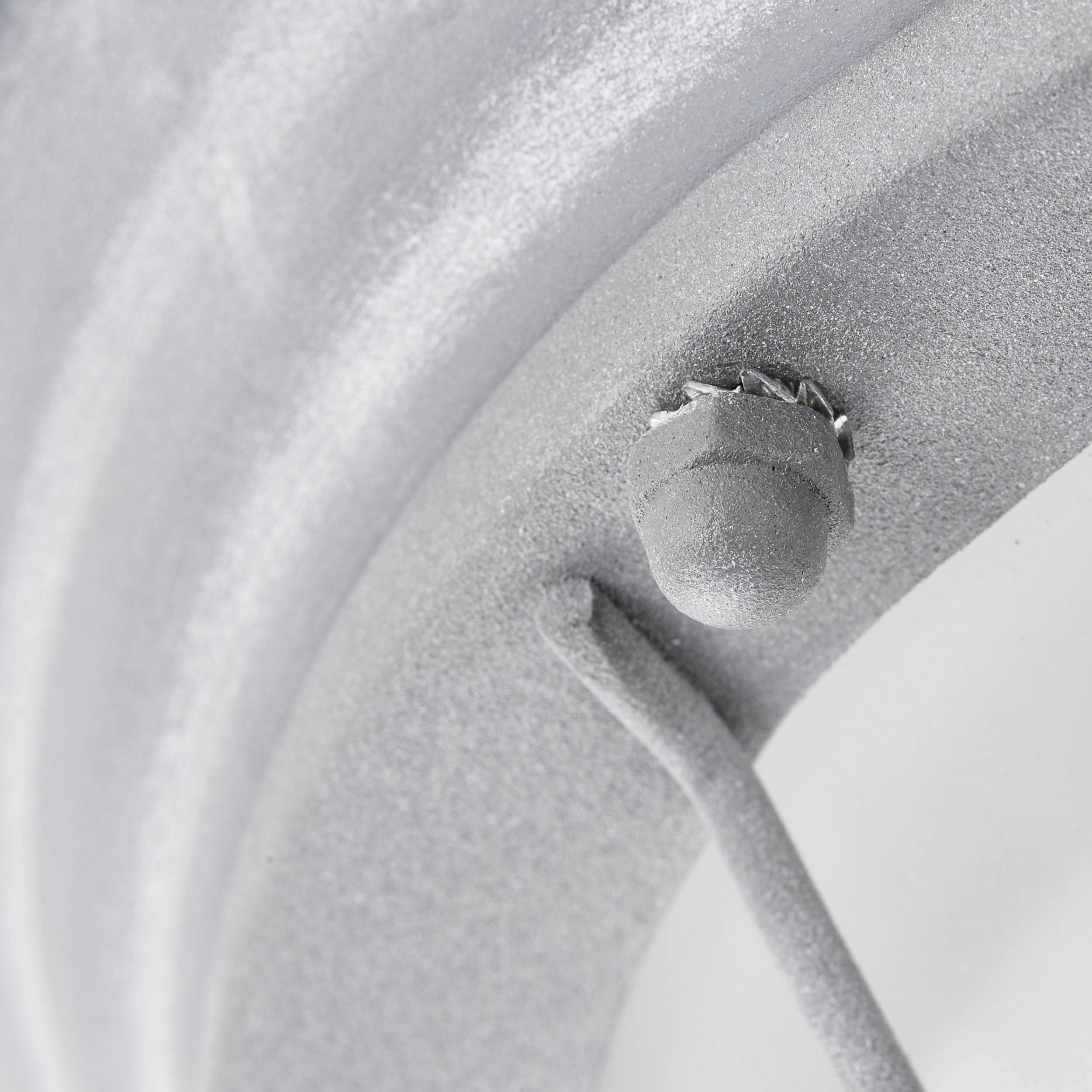 Detailný záber strieborného kovového predmetu s oblými tvarmi a malým vyvýšeným detailom v pravom hornom rohu.