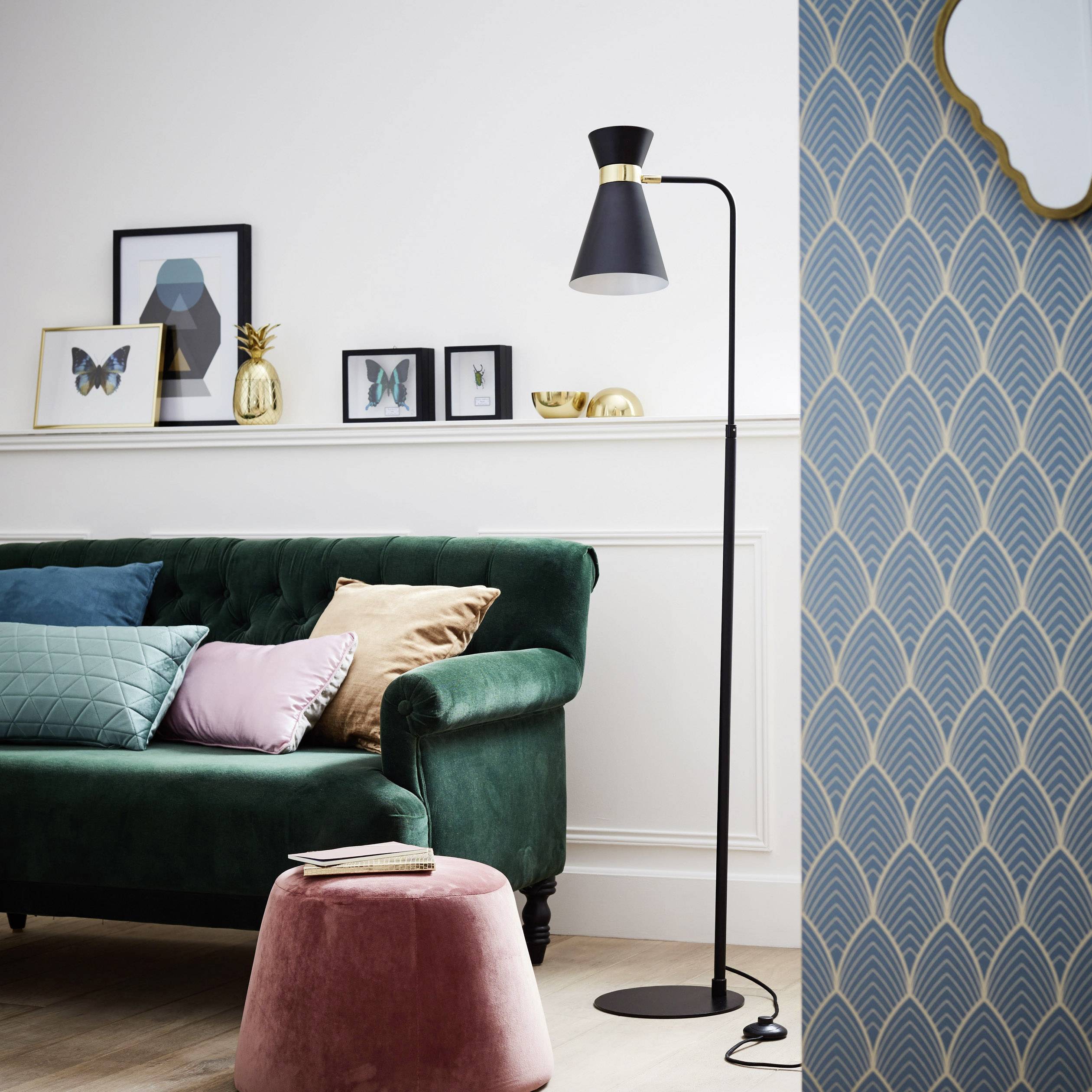Štýlovo zariadená obývačka so zelenou pohovkou, ružovou taburetkou a čiernou stojanovou lampou pred vzorovanou tapetou.