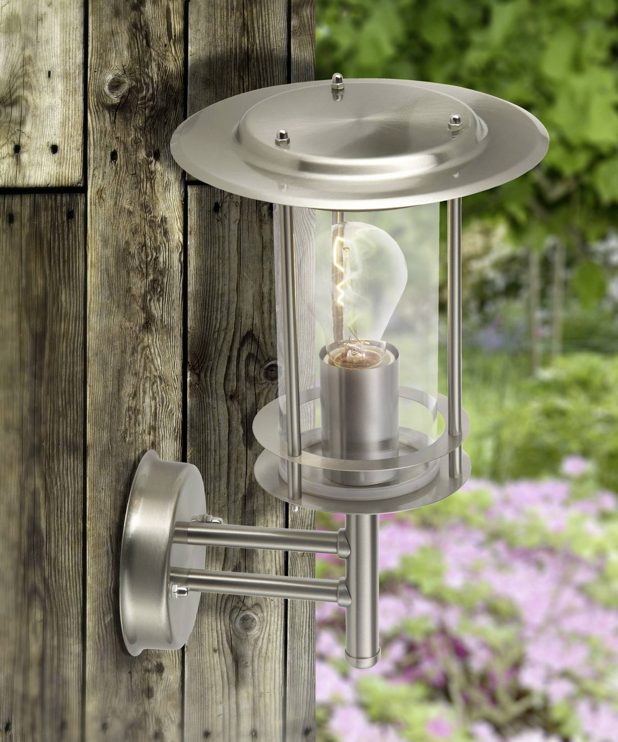 Strieborná vonkajšia nástenná lampa z kovu a skla, namontovaná na drevenej stene. V pozadí sú rozmazané rastliny.