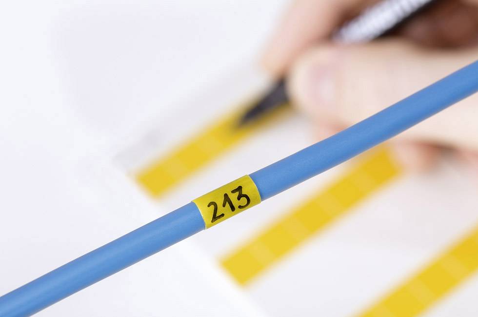 Modré kábel so žltou nálepkou '243', rozmazaná ruka v pozadí, ktorá upravuje kontrolný zoznam so žltými zvýrazneniami.