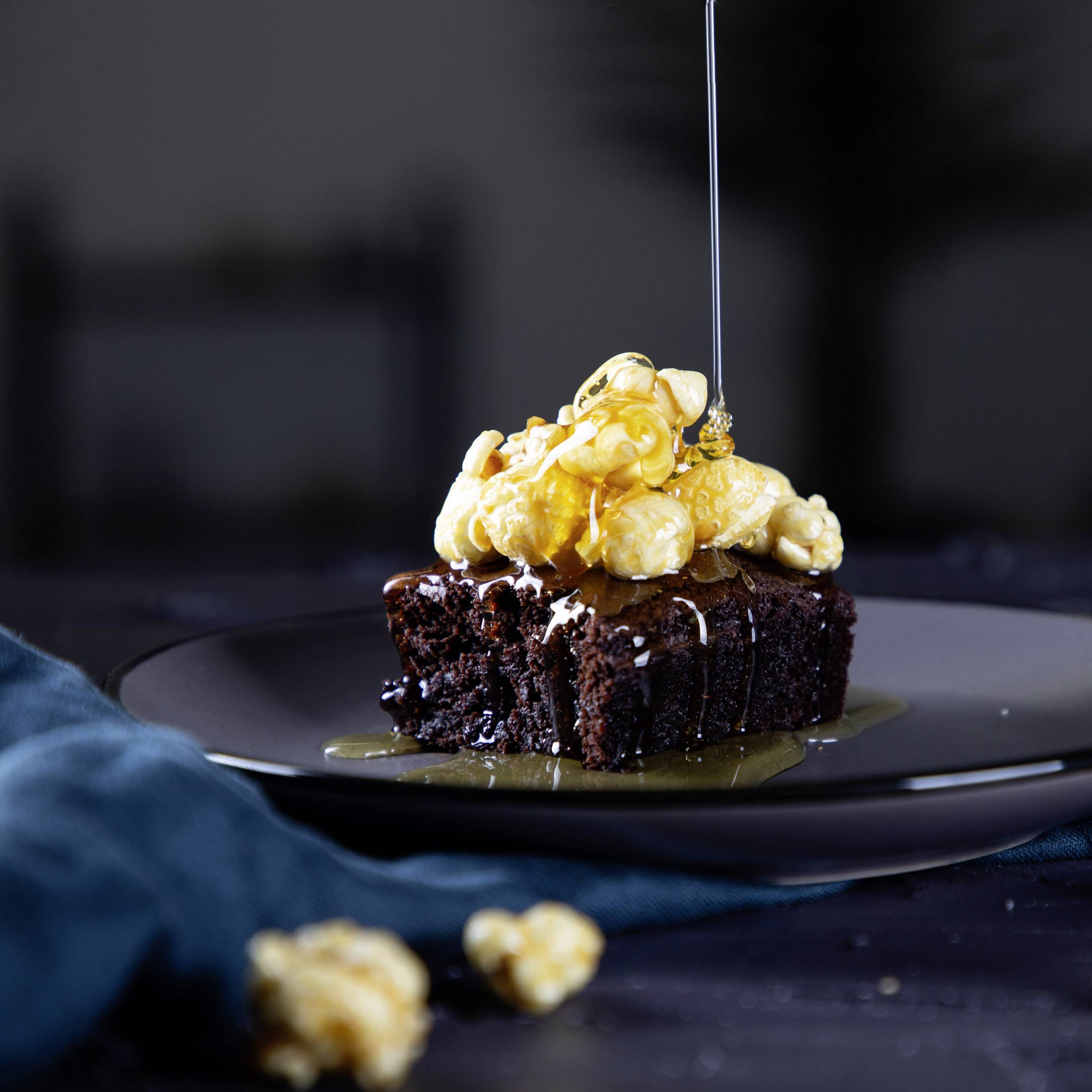 Kúsok čokoládovej torty s karamelovou popcornou a kvapkami tekutého karamelu, servírovaný na tmavom tanieri.